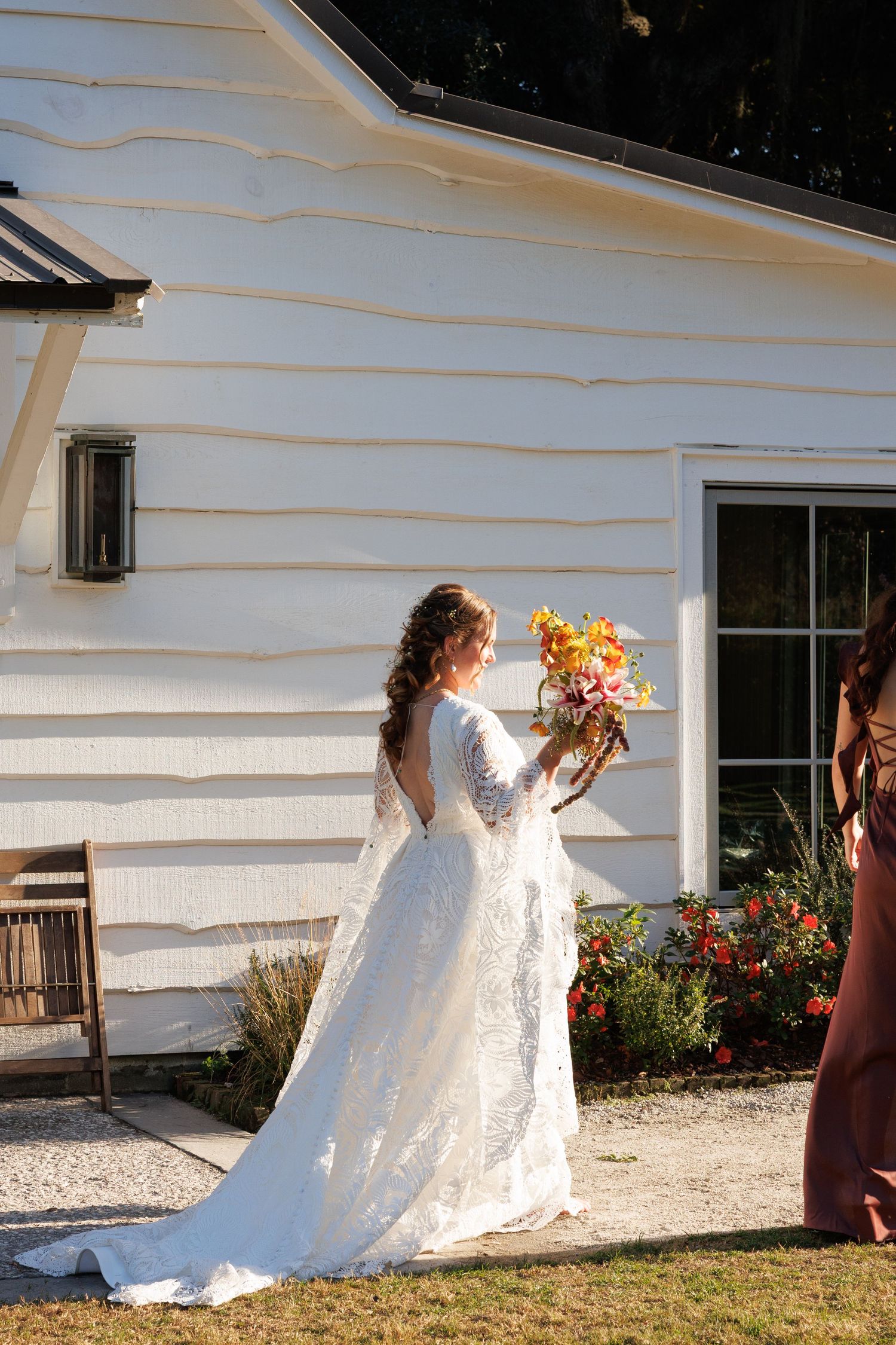 Bride in elegant backless lace wedding gown holding fall bouquet beside white farmhouse building.