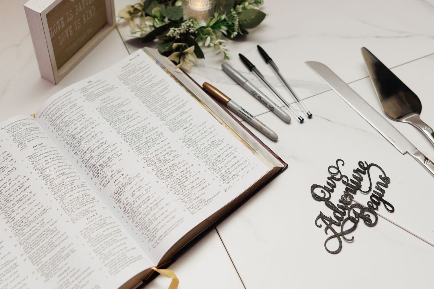 Open Bible and elegant calligraphy sign on a white wedding table with cake serving utensils and greenery decor.