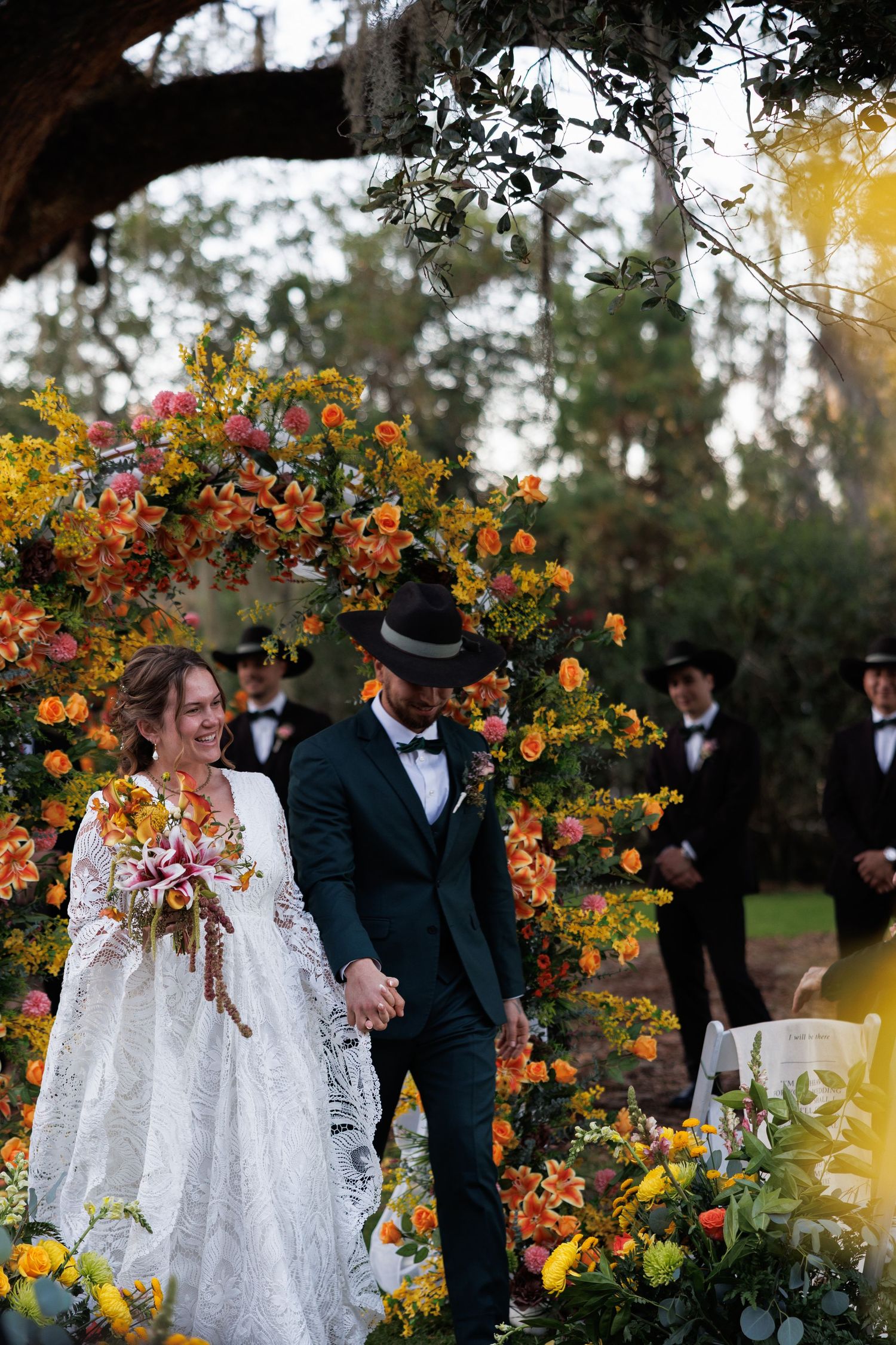 Bride and groom walk hand in hand through vibrant orange and yellow floral wedding arch outdoors.