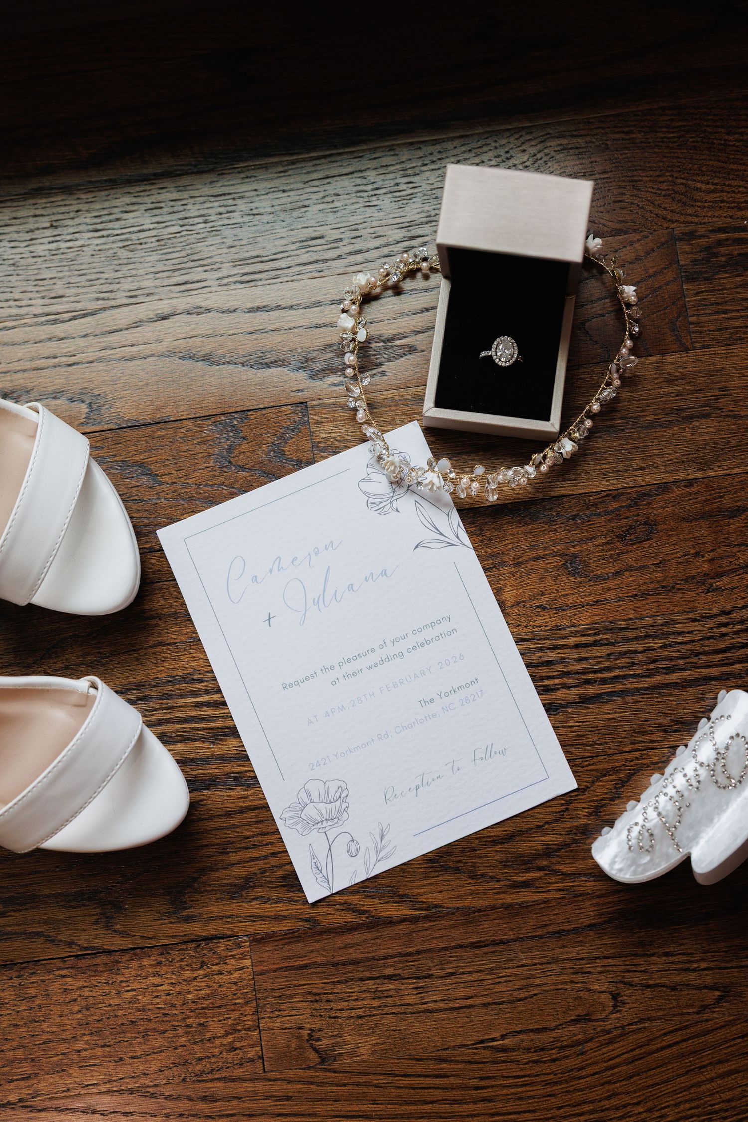 Flat lay of wedding invitation, diamond ring in box, pearl hair piece, white heels, and bridal accessories.