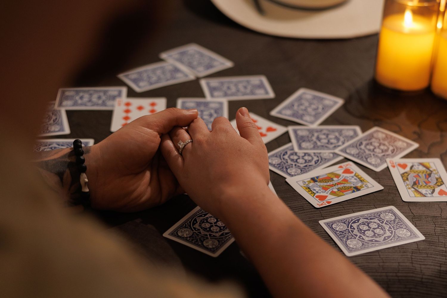 Hands with rings rest on a wooden table surrounded by spread playing cards and glowing candles in dim light.