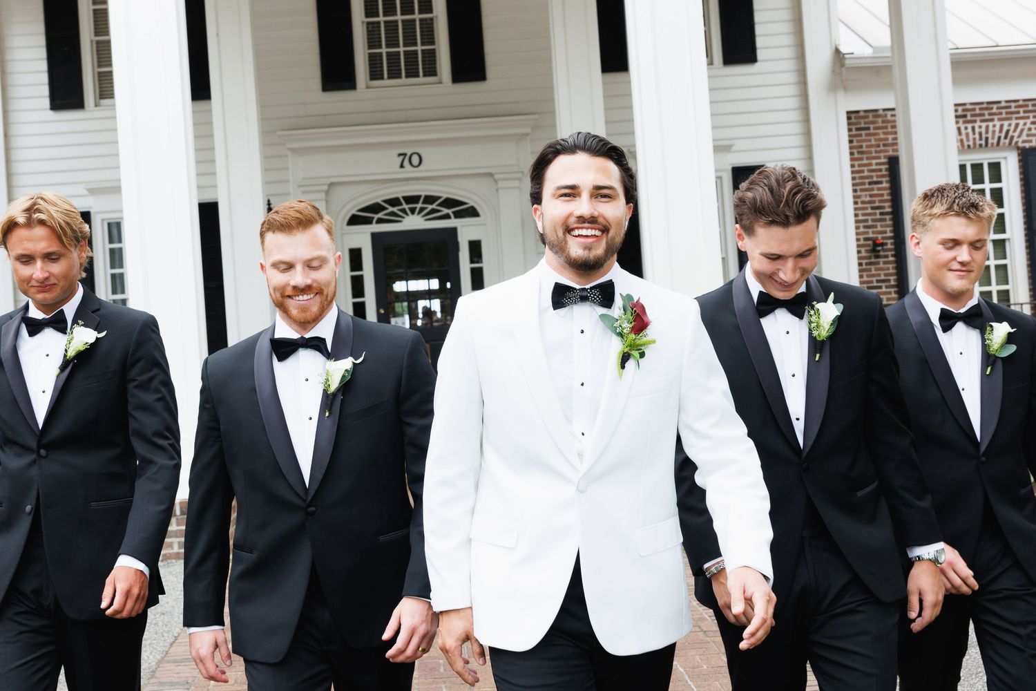 Groom in white tuxedo jacket walks with groomsmen in black suits outside a white colonial building.
