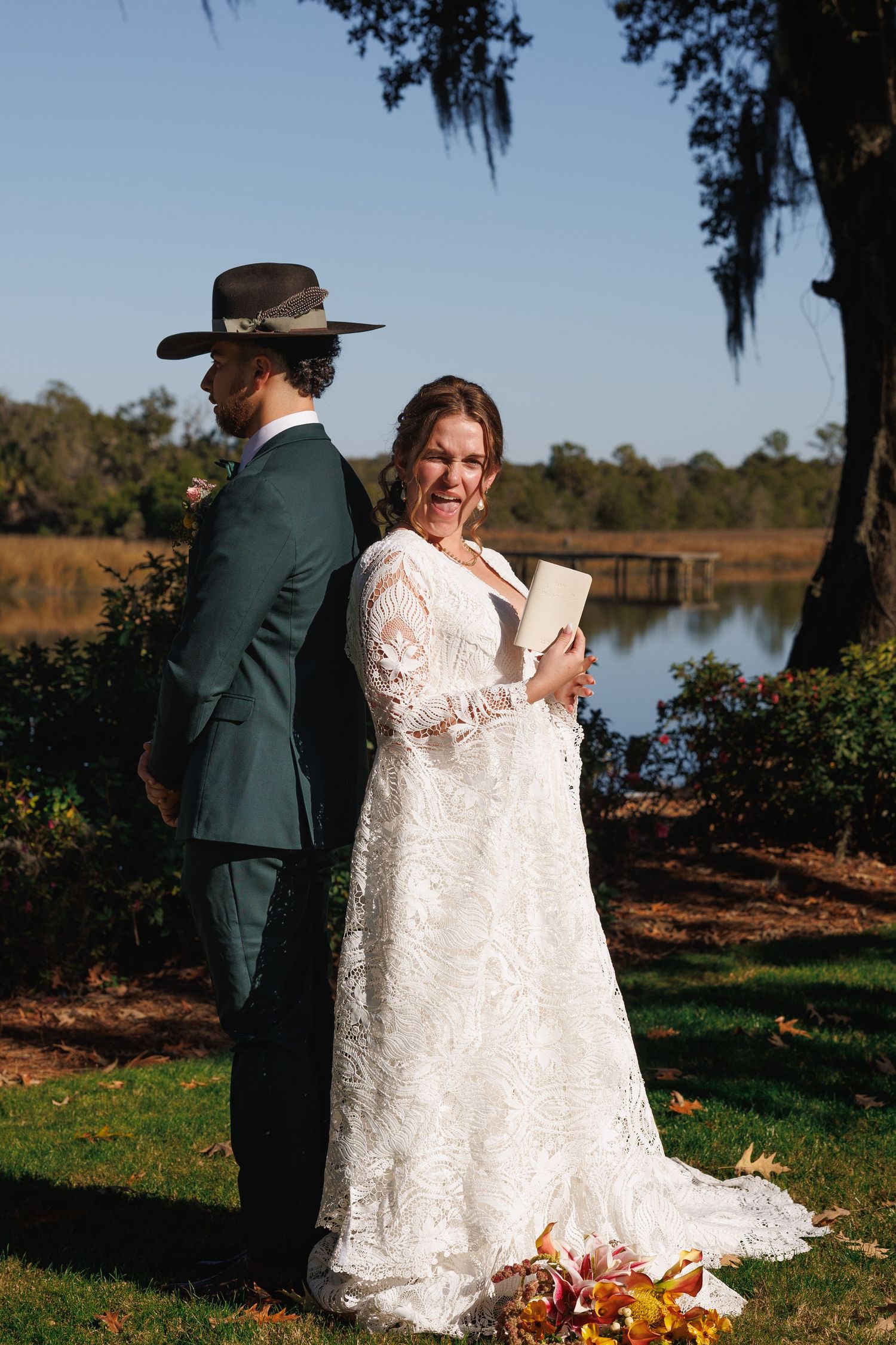 Bride in lace gown reads vows while groom stands back-to-back with her near a sunlit lake.