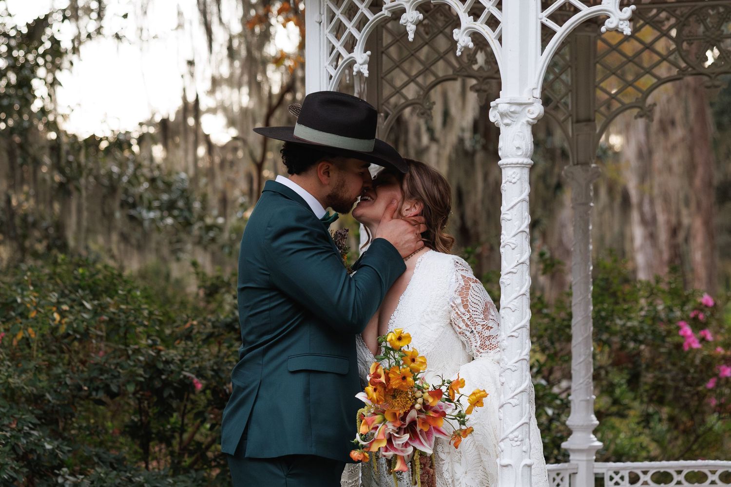 Bride and groom share a kiss under a white ornate gazebo with Spanish moss trees behind them.
