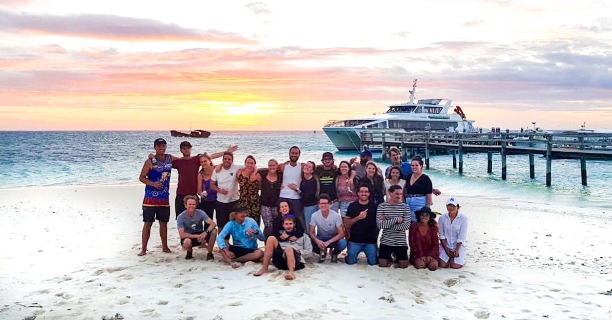 Group of tourists posing on a tropical beach at sunset with a cruise ship docked at a pier in the background.