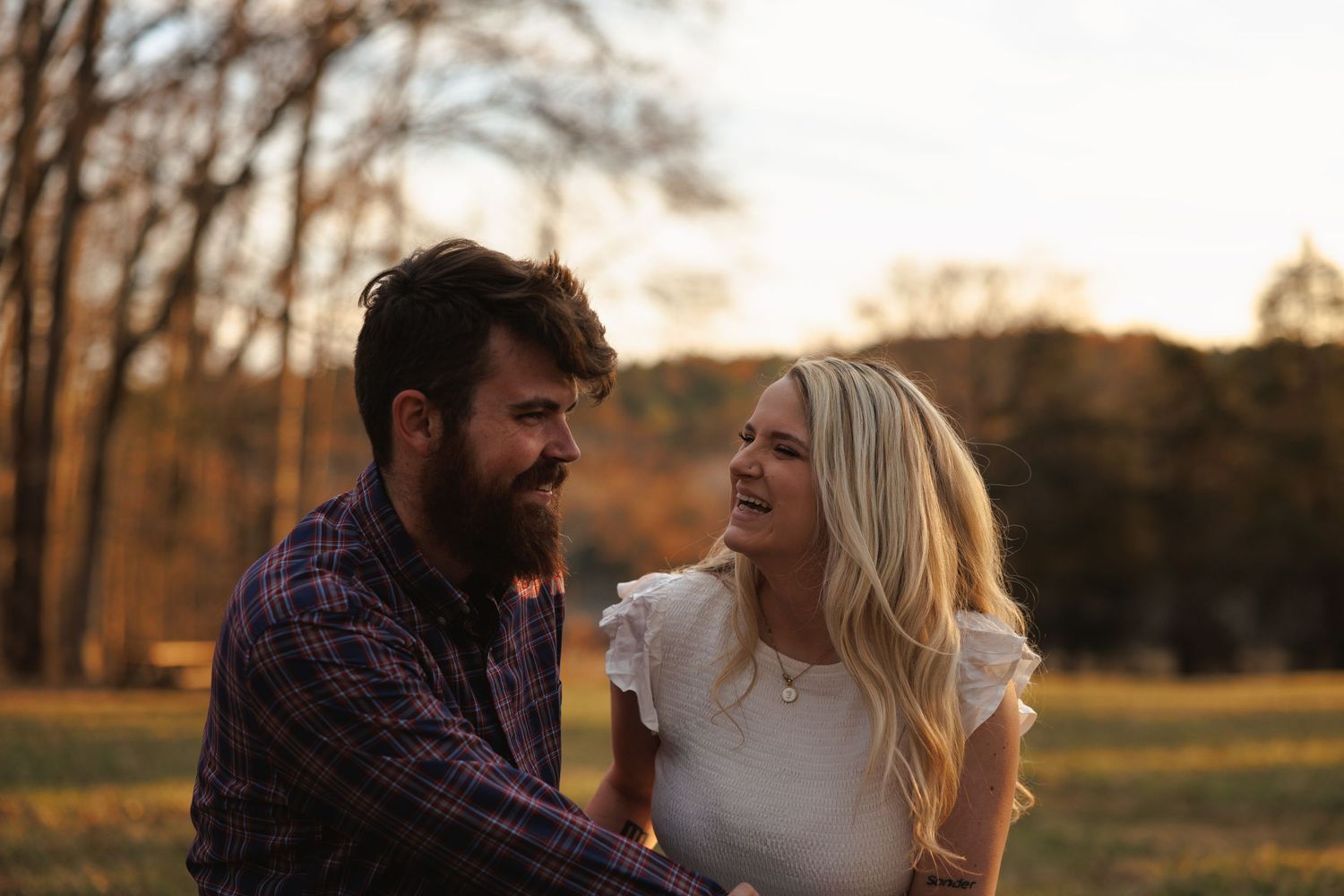 A bearded man and smiling blonde woman share a tender moment together in a sunlit autumn field.