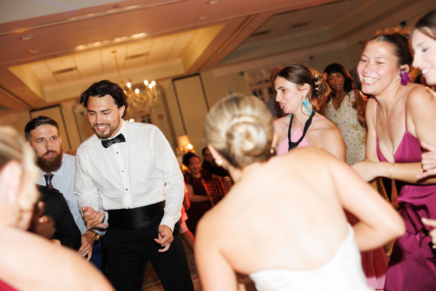 Wedding guests dance joyfully with groom in tuxedo and bride in white dress at reception.
