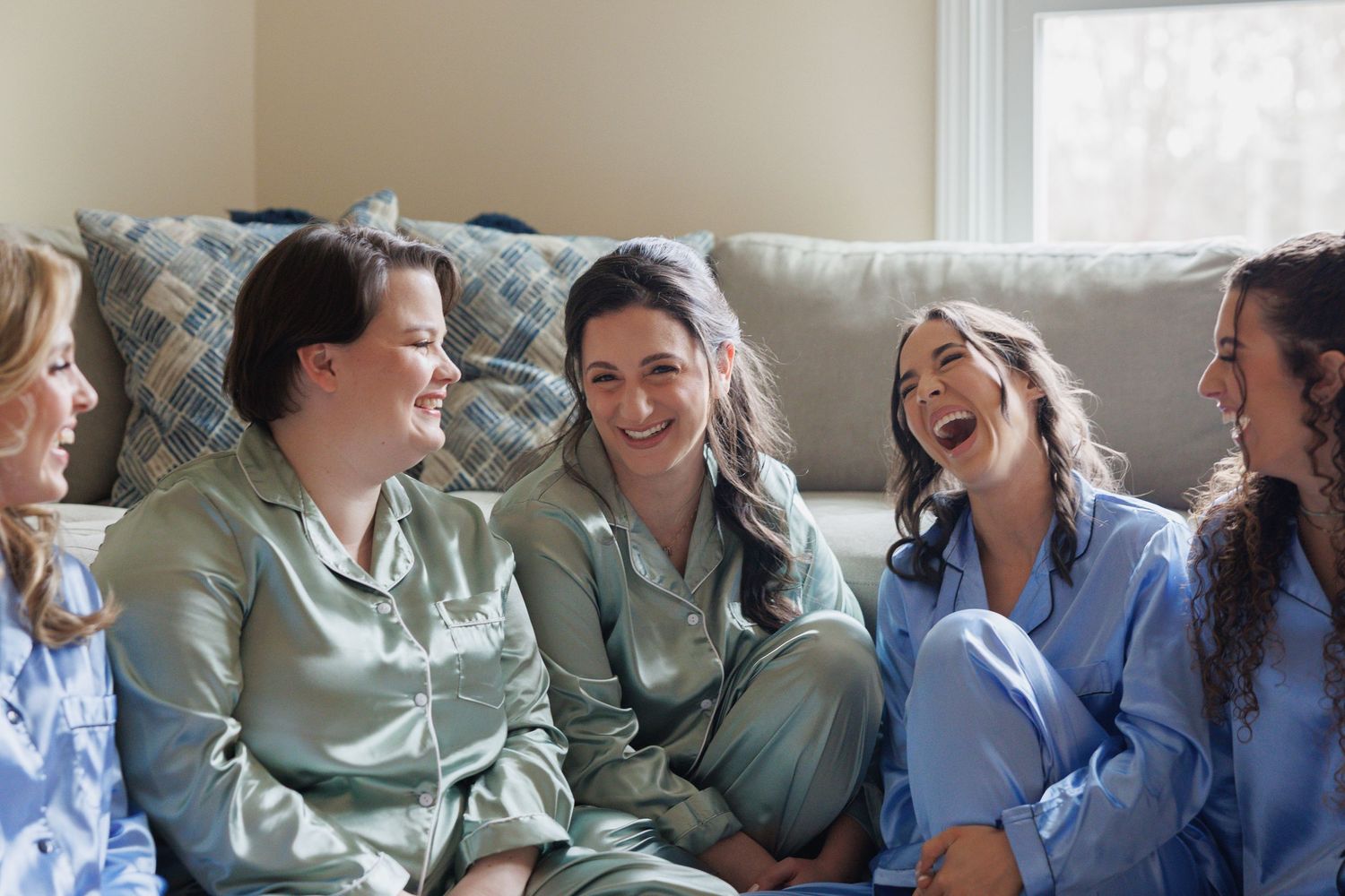 Five women in satin pajamas laughing and bonding together during a fun sleepover or bachelorette party gathering.