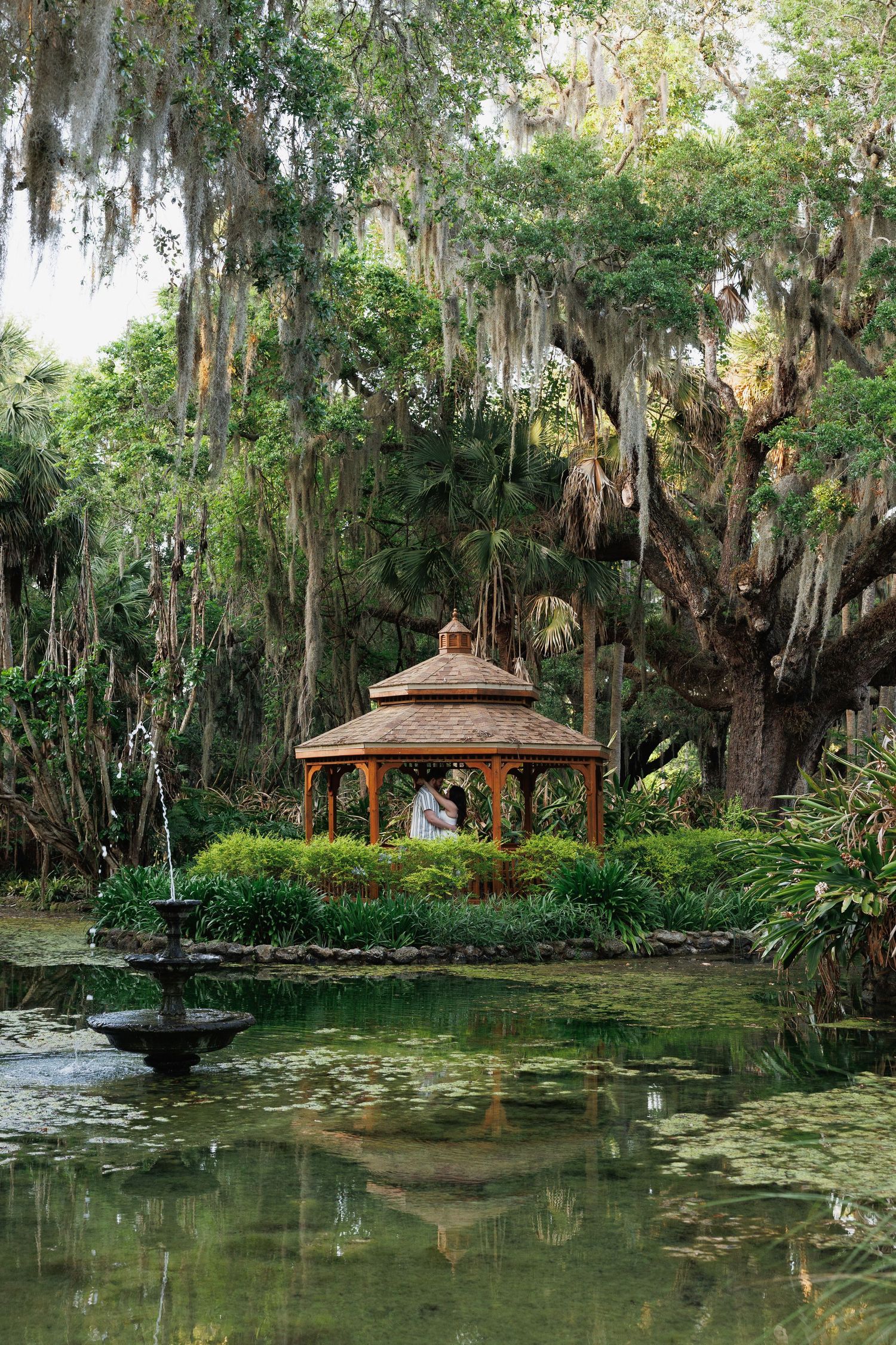 A couple shares a romantic moment in a wooden gazebo on a serene pond with Spanish moss-draped oak trees.