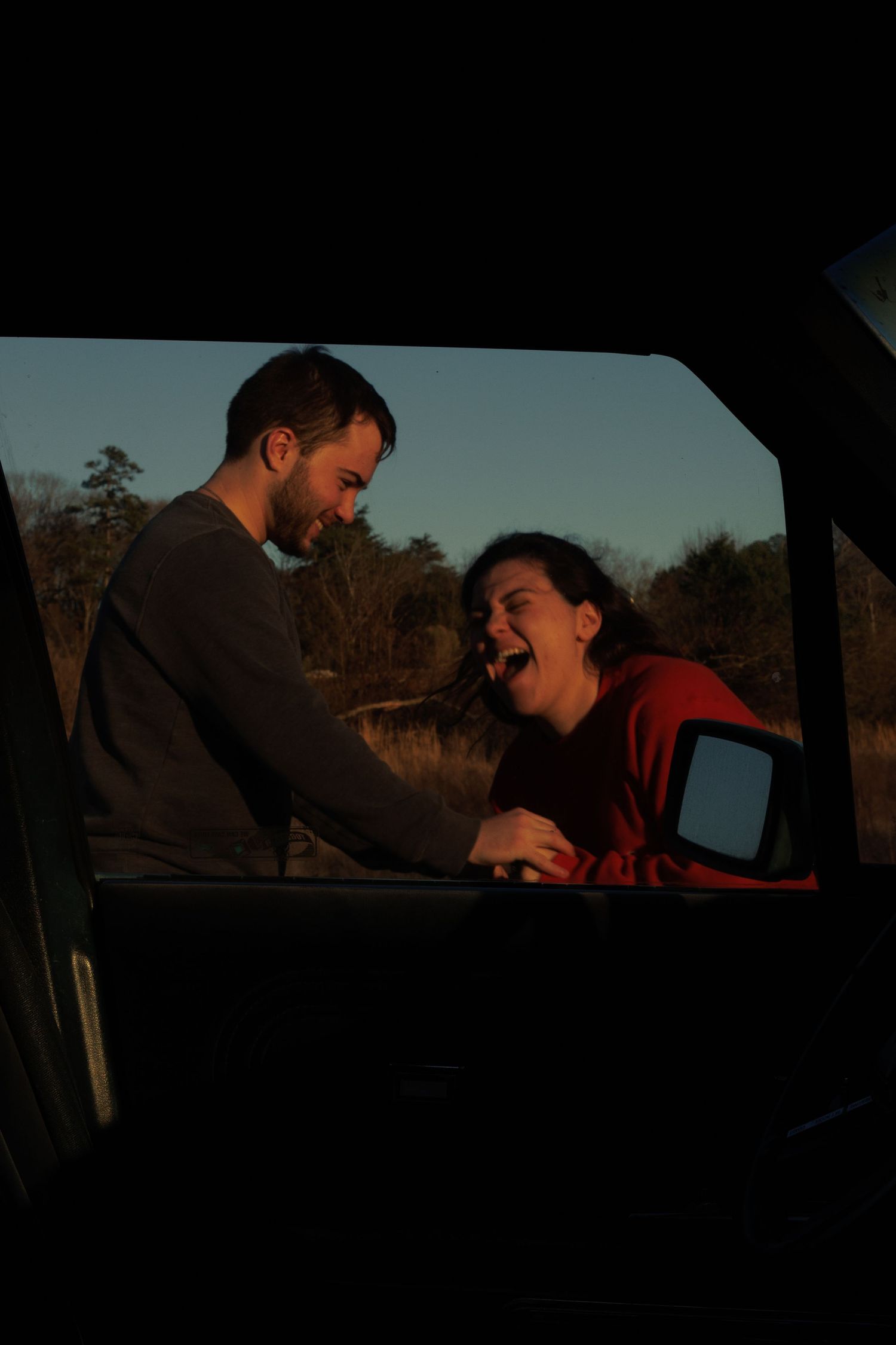 A laughing woman in red and a smiling man share a joyful moment viewed through a car window at dusk.