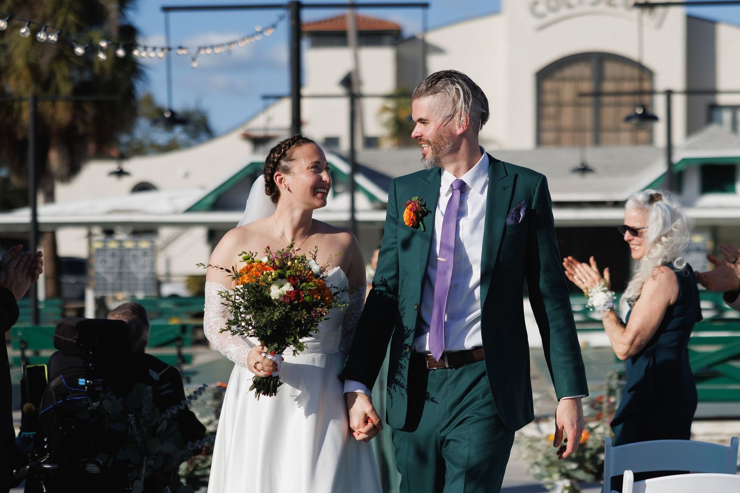 Joyful newlywed couple walks down the aisle holding hands, bride carrying colorful bouquet at outdoor wedding ceremony.
