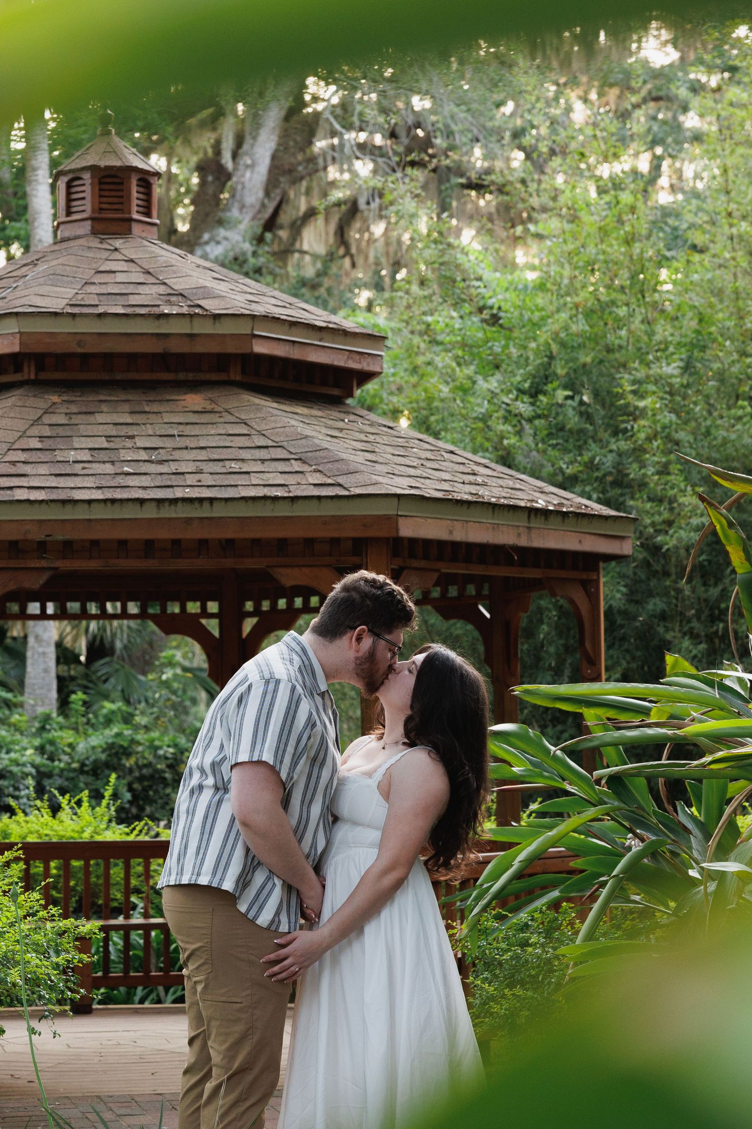 Couple sharing a romantic kiss in front of a wooden gazebo surrounded by lush green tropical gardens.