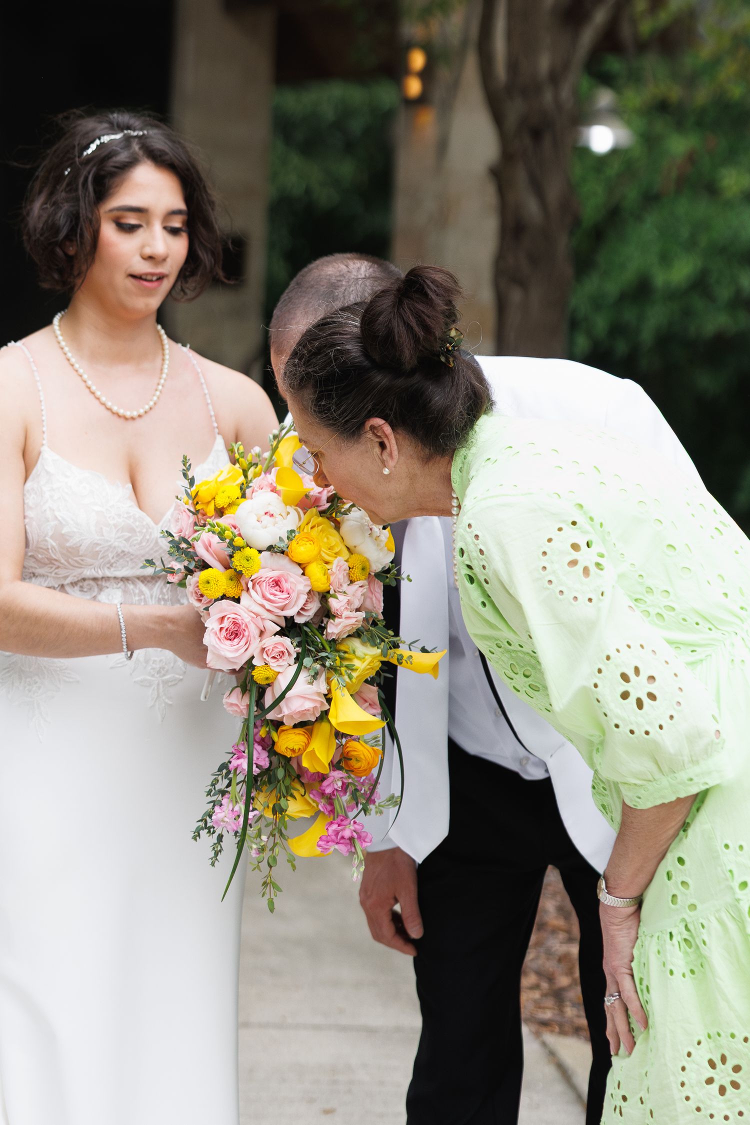 Bride in white gown holding colorful yellow and pink bouquet while guest leans in to smell flowers.