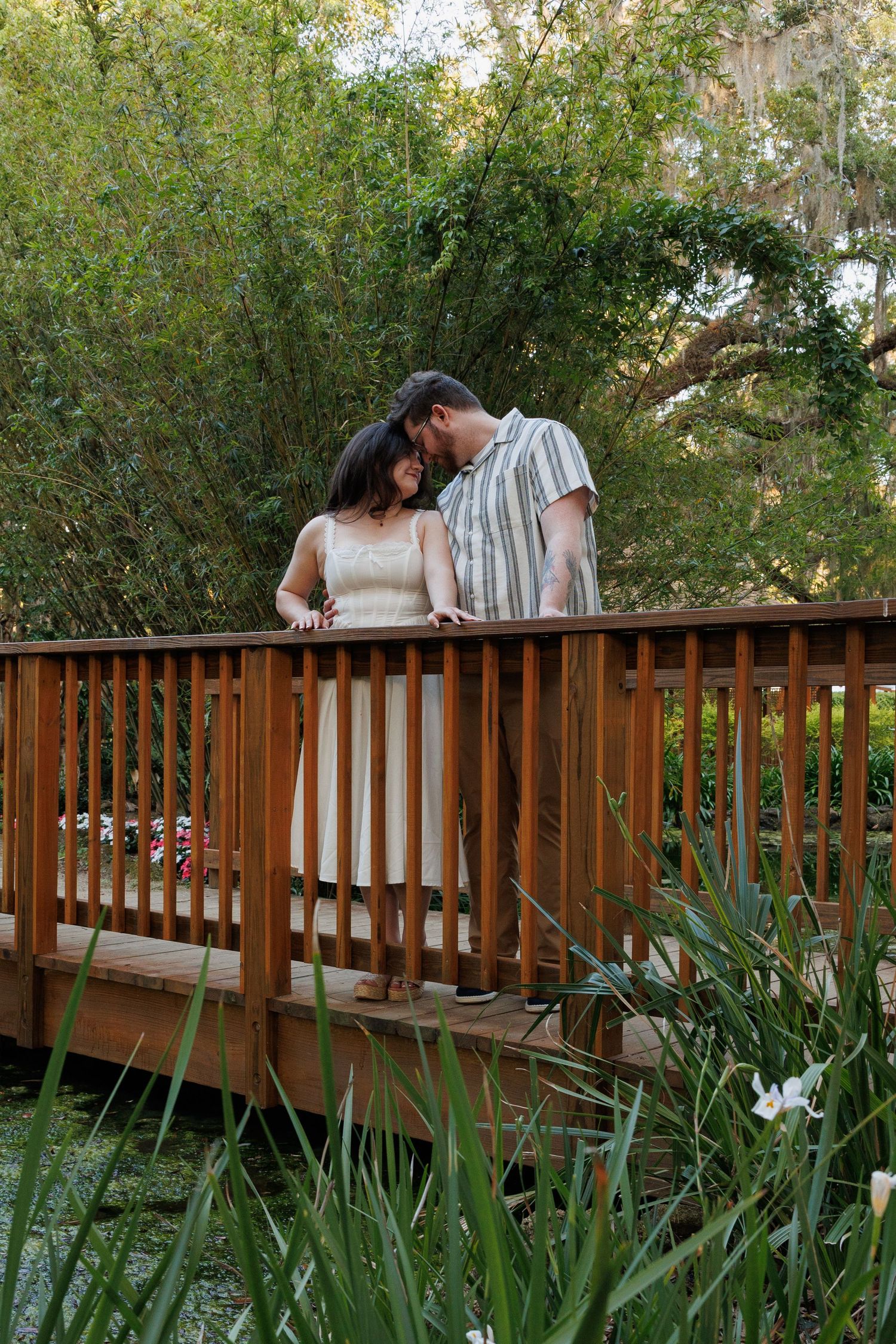 Couple sharing a romantic moment on a wooden bridge surrounded by lush greenery and pond.