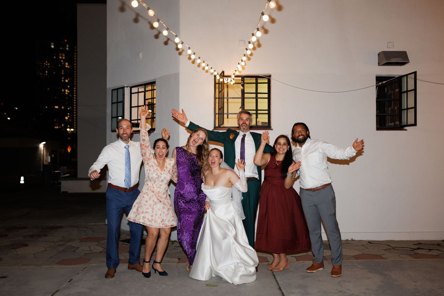 Wedding party group posing joyfully outdoors at night with string lights during reception celebration.