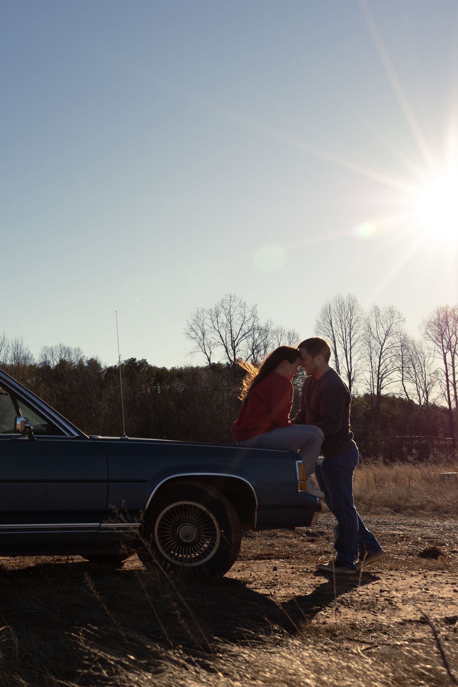 Couple leaning in for a kiss against a classic blue car with sun flare in an open field.