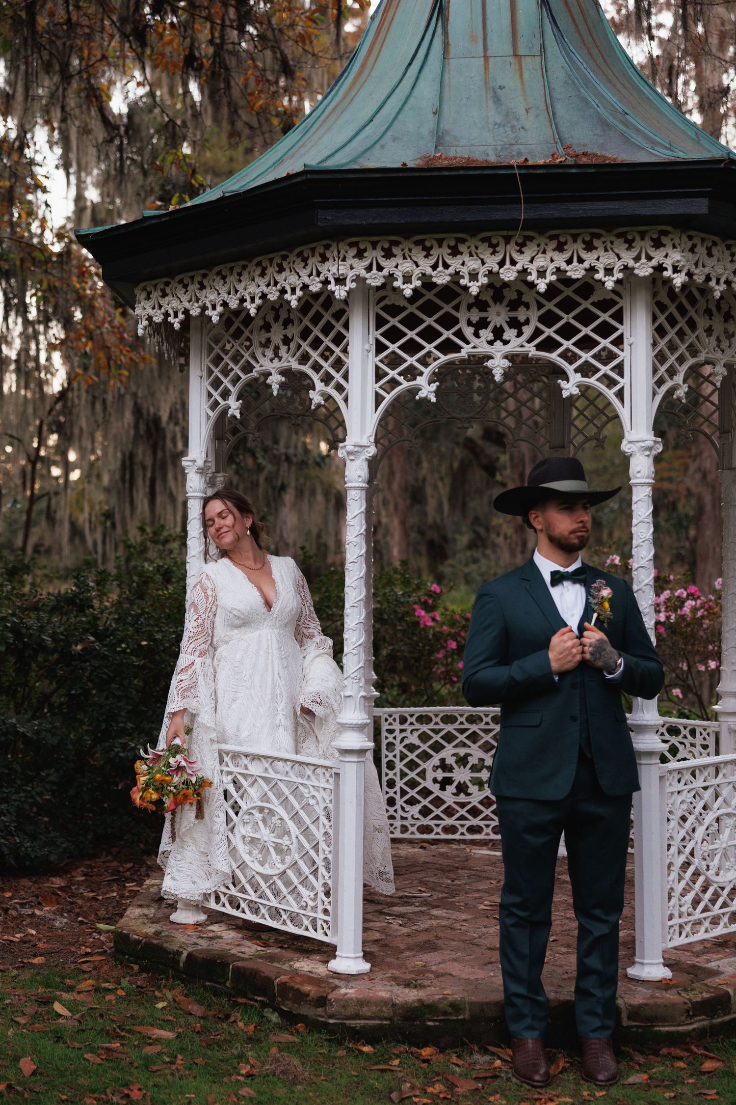 Bride in lace gown and groom in teal suit with cowboy hat pose at a white Victorian gazebo.