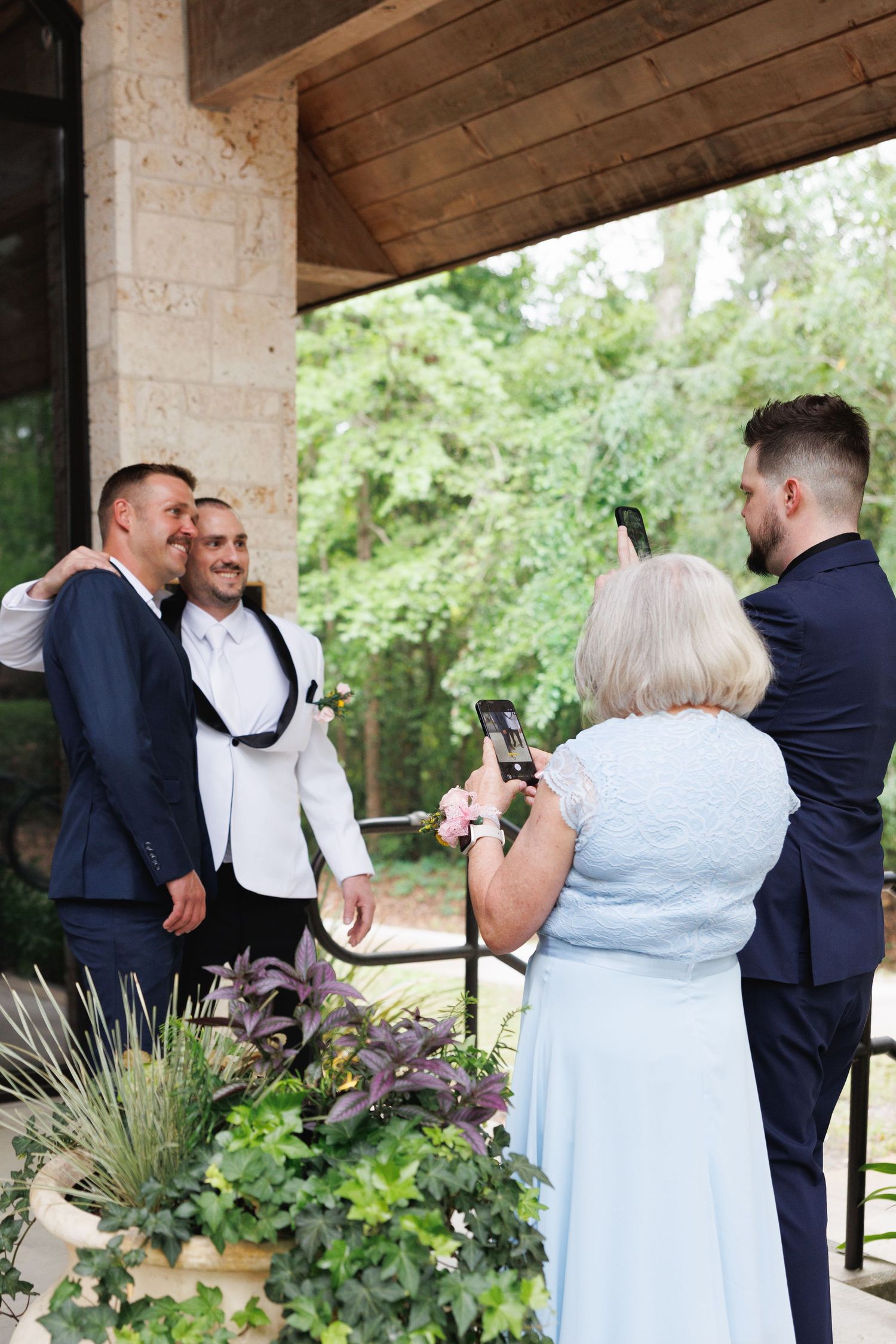 Two grooms pose for photos with family members capturing the moment on smartphones at an outdoor wedding venue.