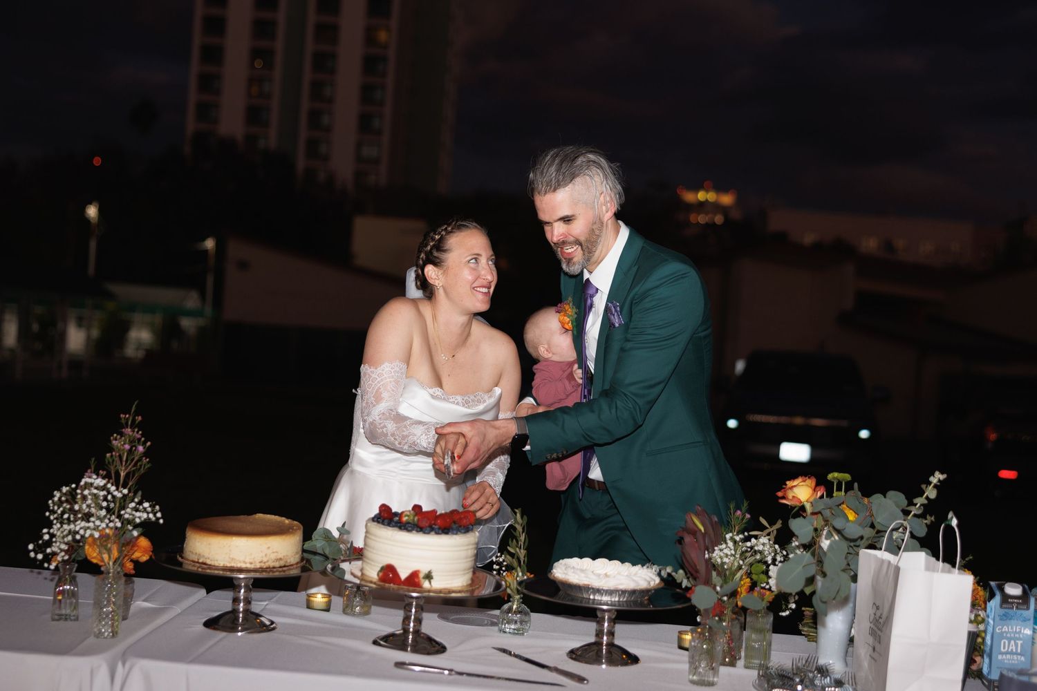 Bride and groom cutting wedding cake together at night with floral arrangements on the table.