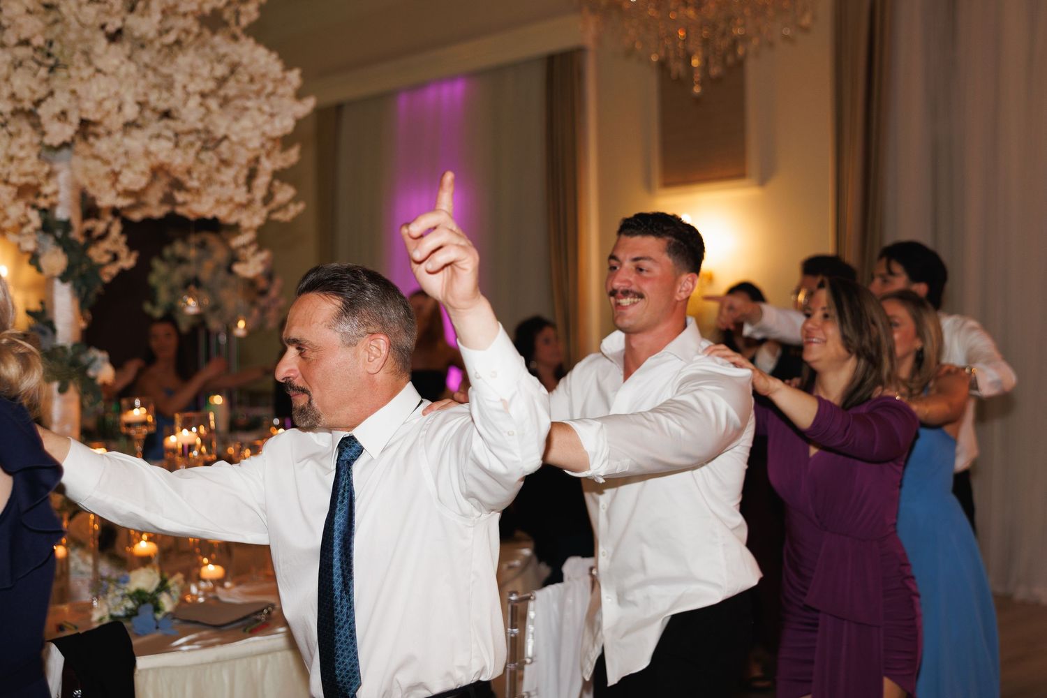 Wedding guests dance joyfully at a reception in an elegant ballroom with floral decorations.