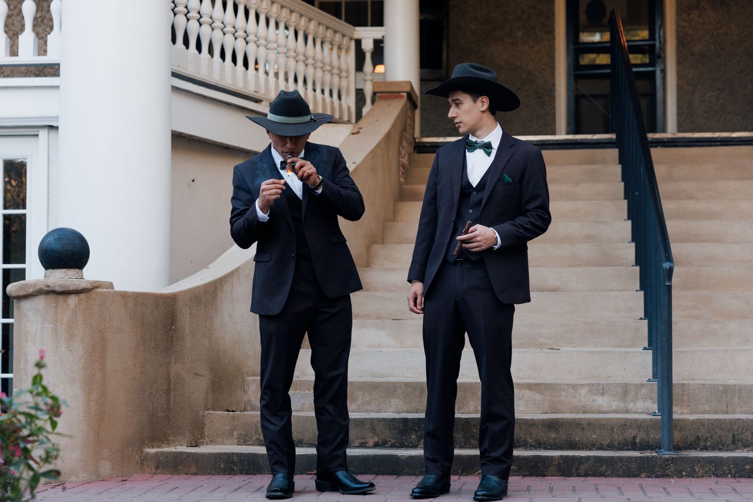 Two men in black suits and cowboy hats standing outside a elegant building before a wedding.