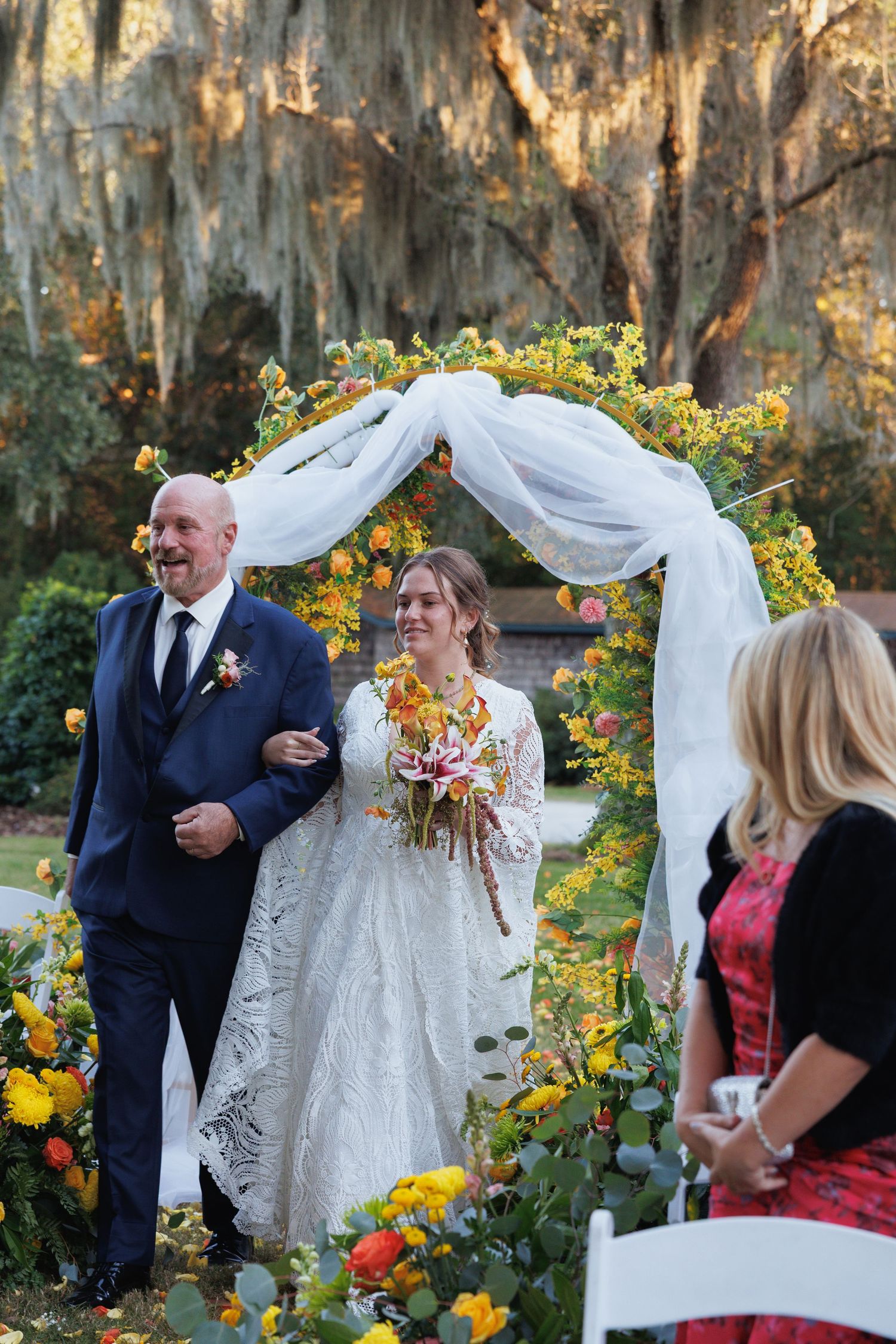 Bride and groom smiling under a floral arch with yellow and orange flowers at an outdoor fall wedding ceremony.
