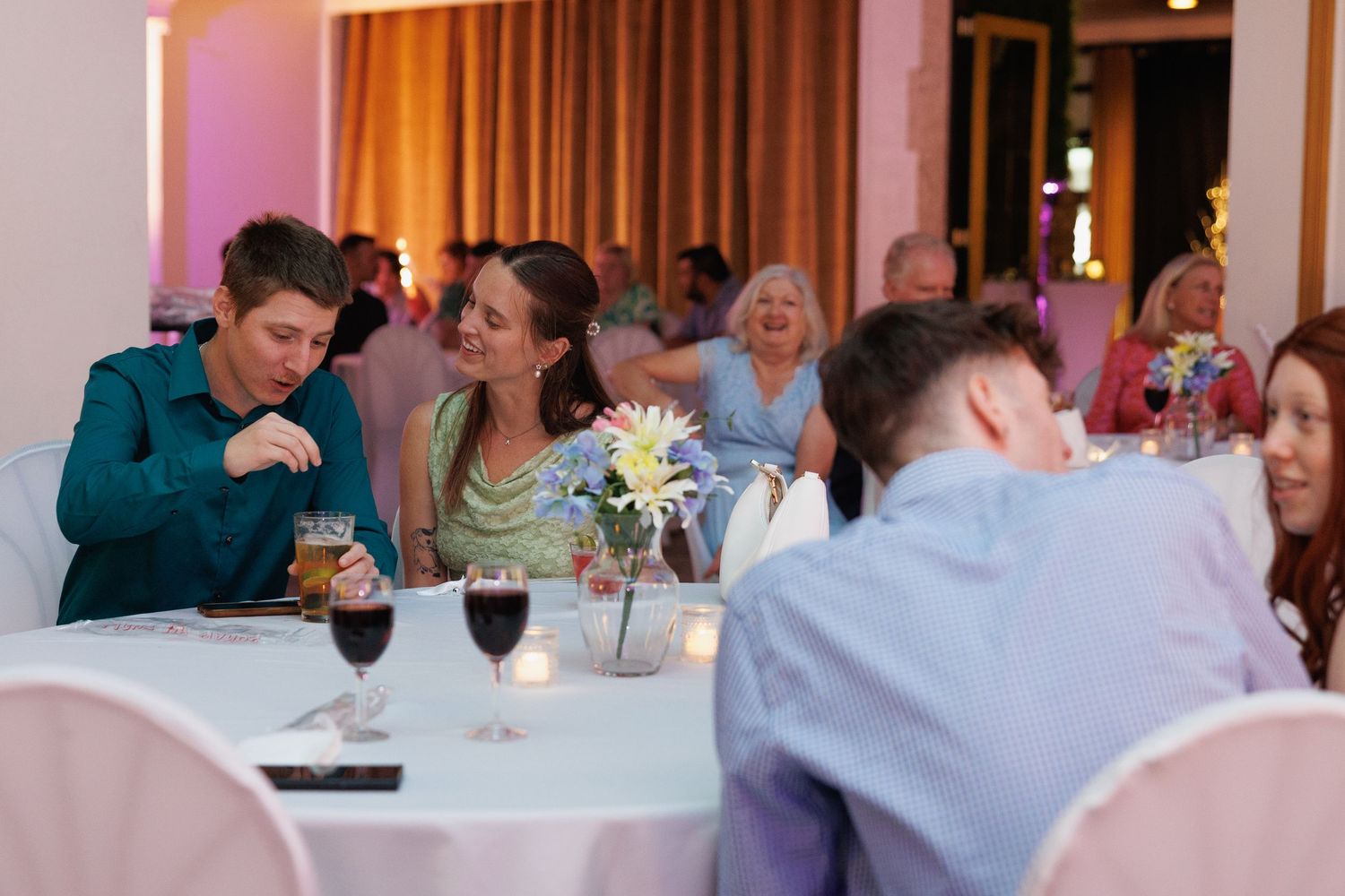 Wedding reception guests dining at round tables with wine glasses in warmly lit ballroom.