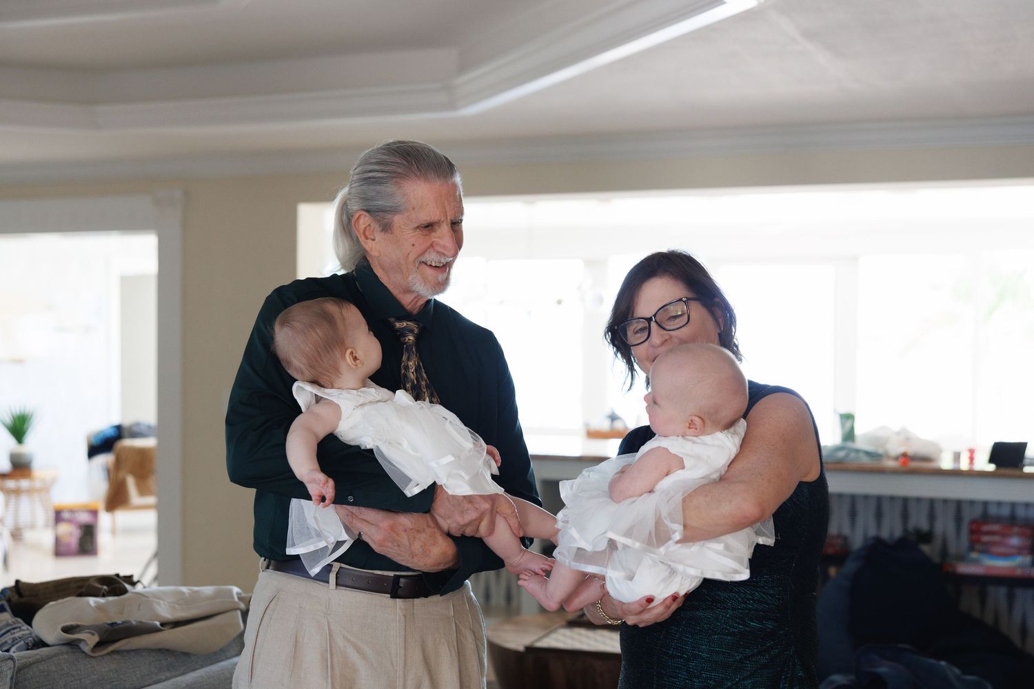 Elderly man and woman with glasses each hold a baby at a bright indoor family gathering celebration.