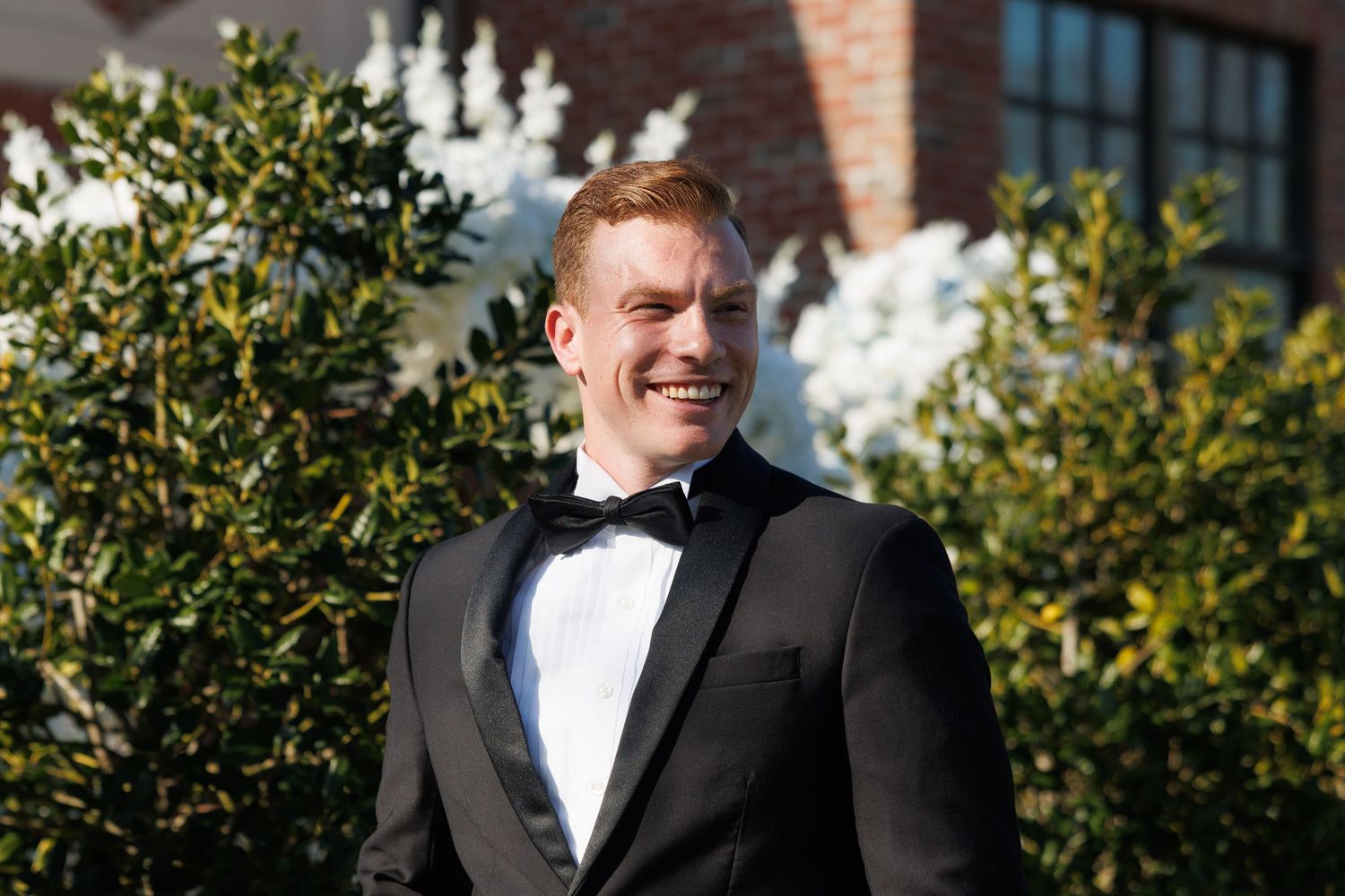 Smiling groom in black tuxedo with bow tie poses outdoors among lush green shrubs.
