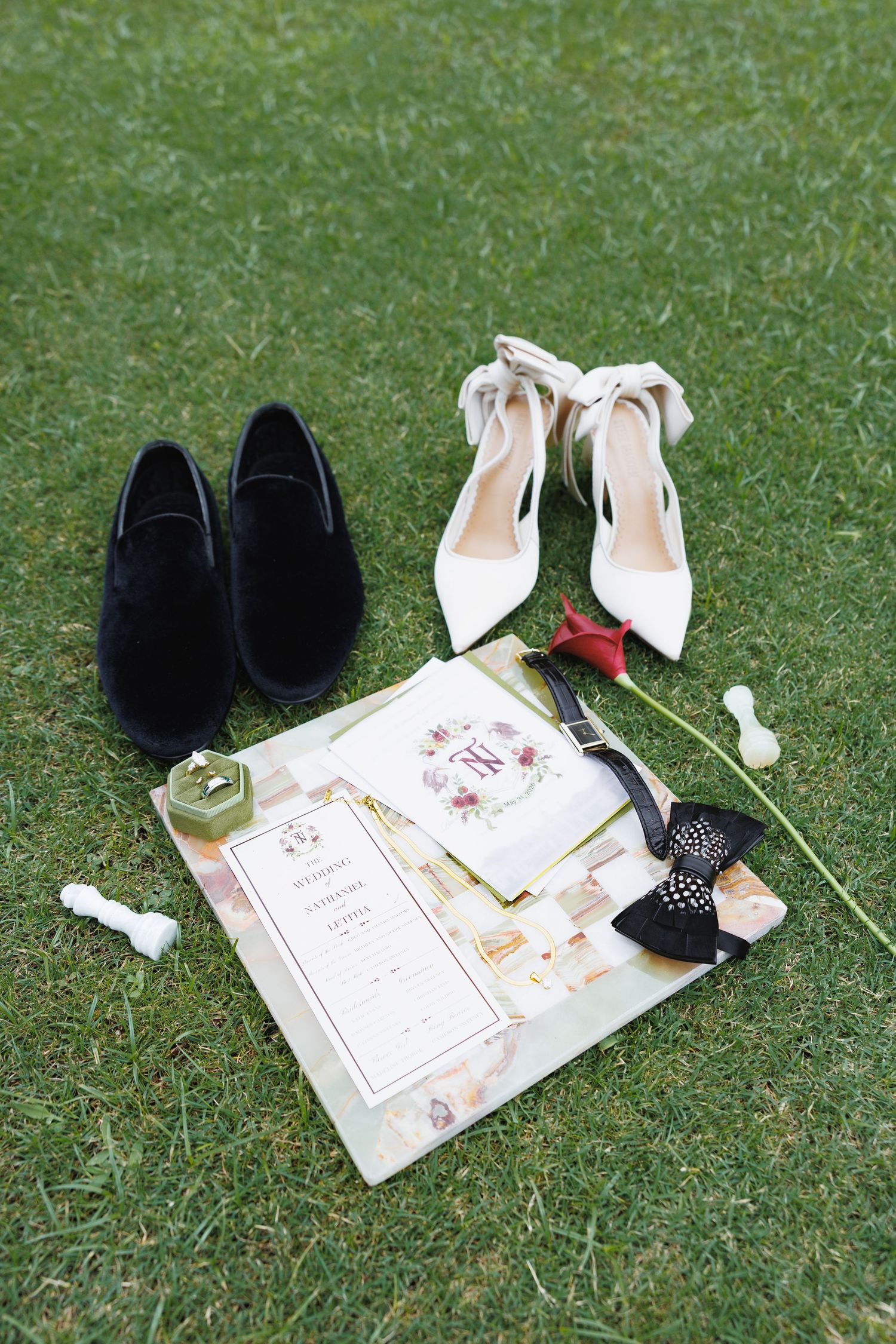 Wedding flat lay on green grass featuring black loafers, white heeled shoes, floral invitations, a red rose, bow tie, and ring box.
