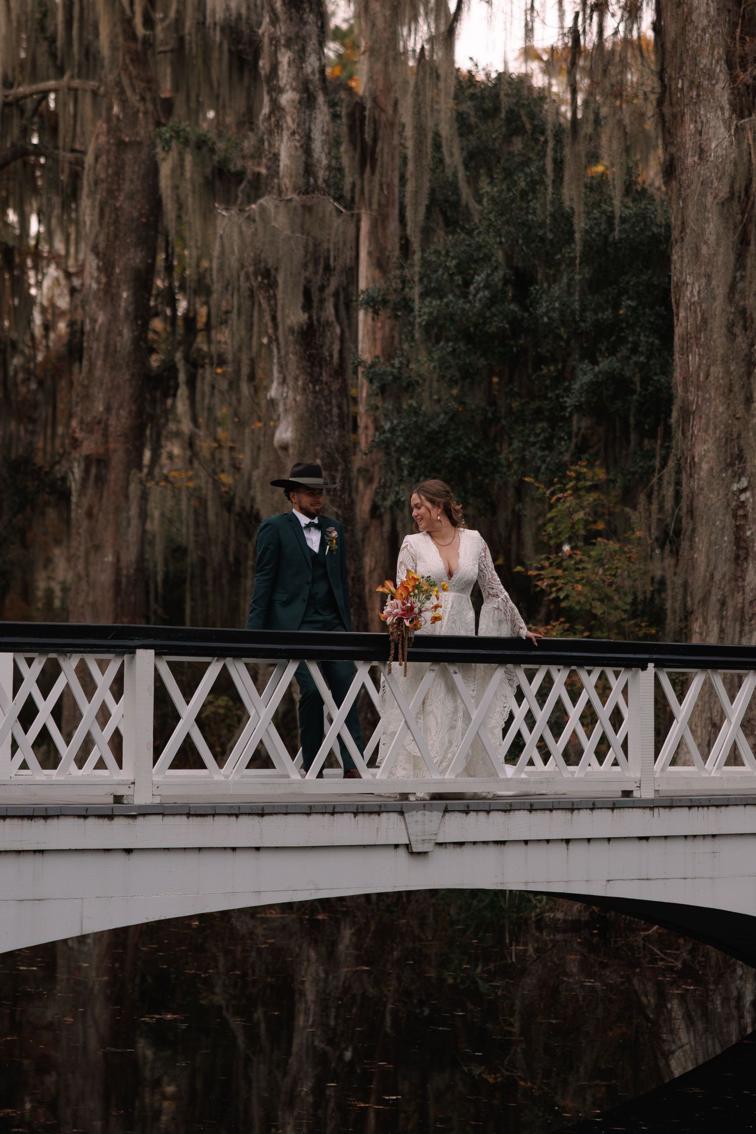 Bride and groom walking on a white ornate bridge surrounded by Spanish moss-draped trees.