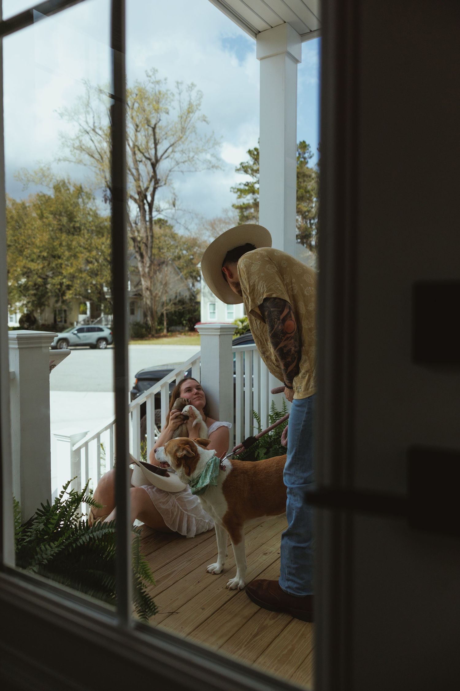 A couple sits on a white porch with their dog, as a tattooed man in a cowboy hat leans over to greet them.