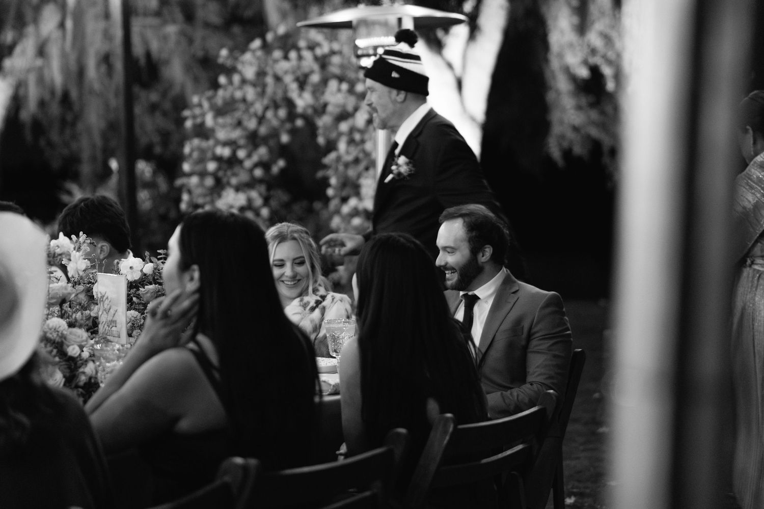 Black and white photo of wedding guests laughing at a reception dinner table outdoors.