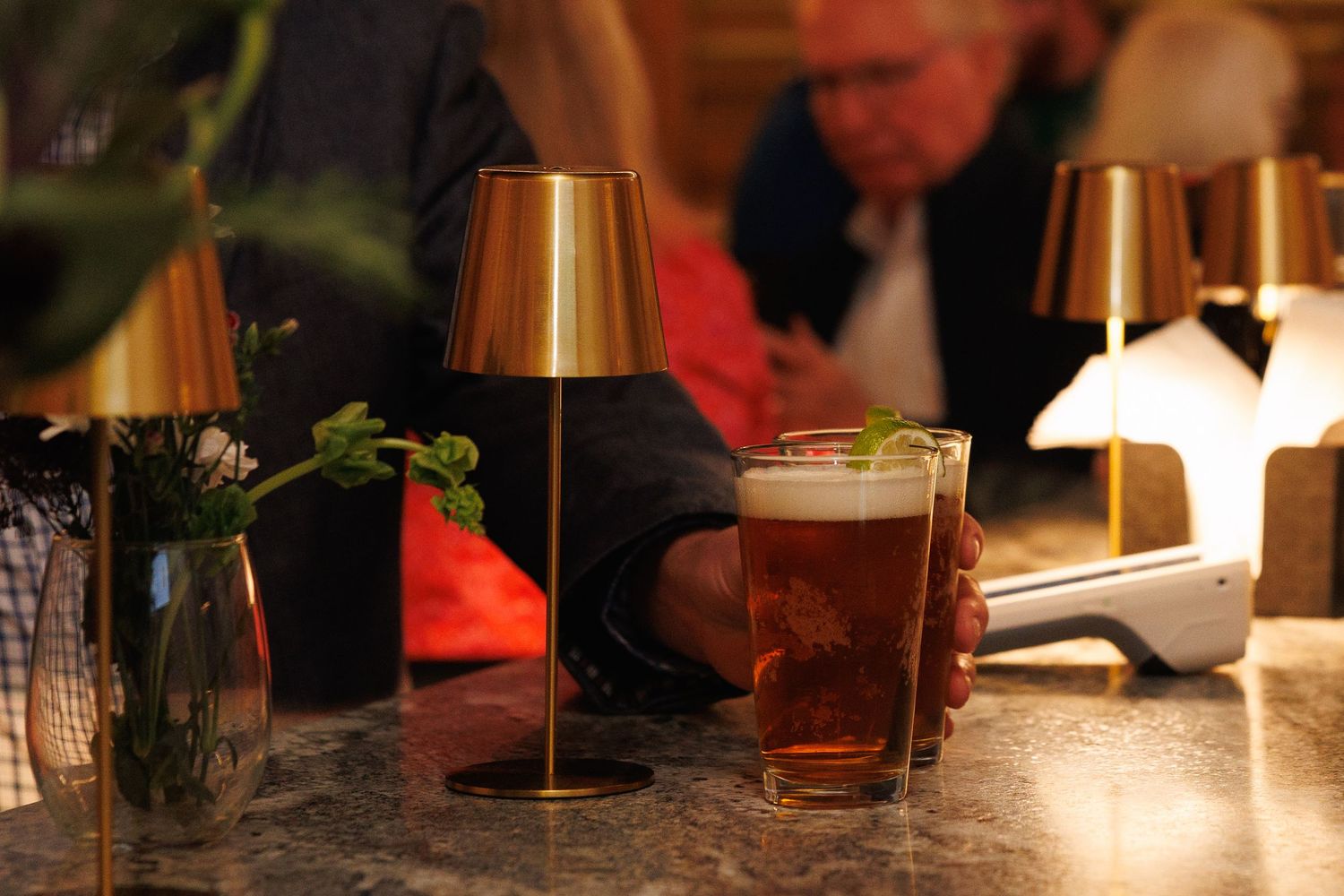 A glass of beer on a bar counter beside a gold lamp, with patrons and warm lighting in the background.