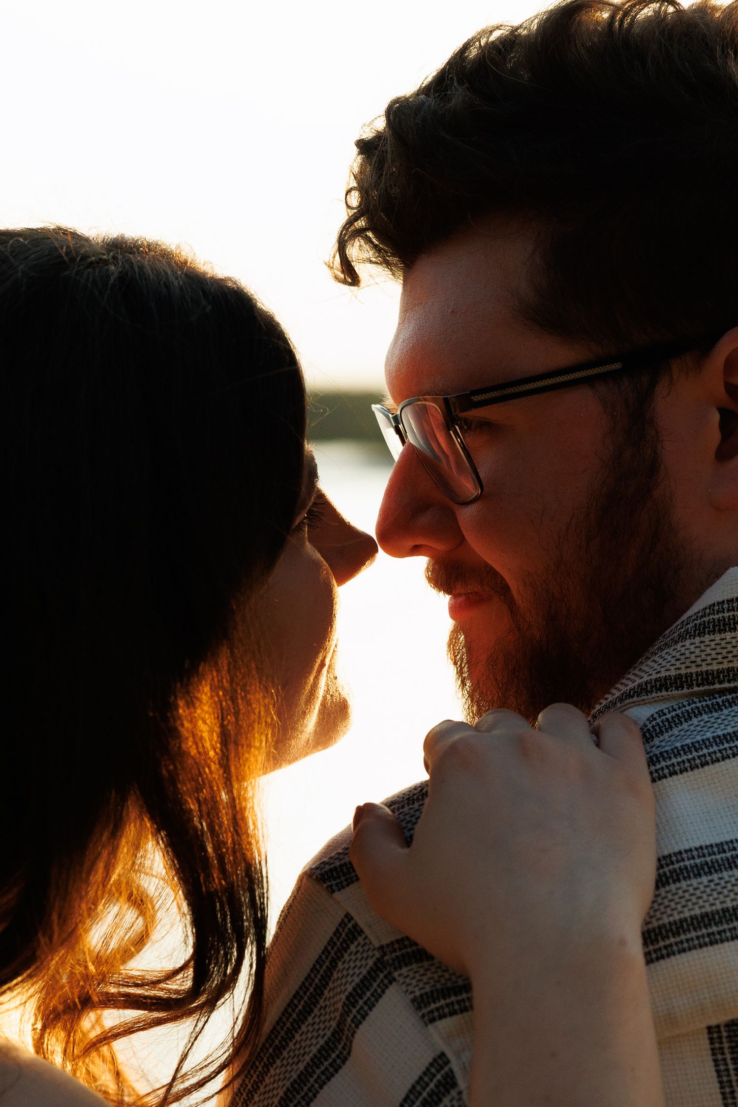 Close-up of a romantic couple nearly kissing at sunset, man wearing glasses and a striped shirt.