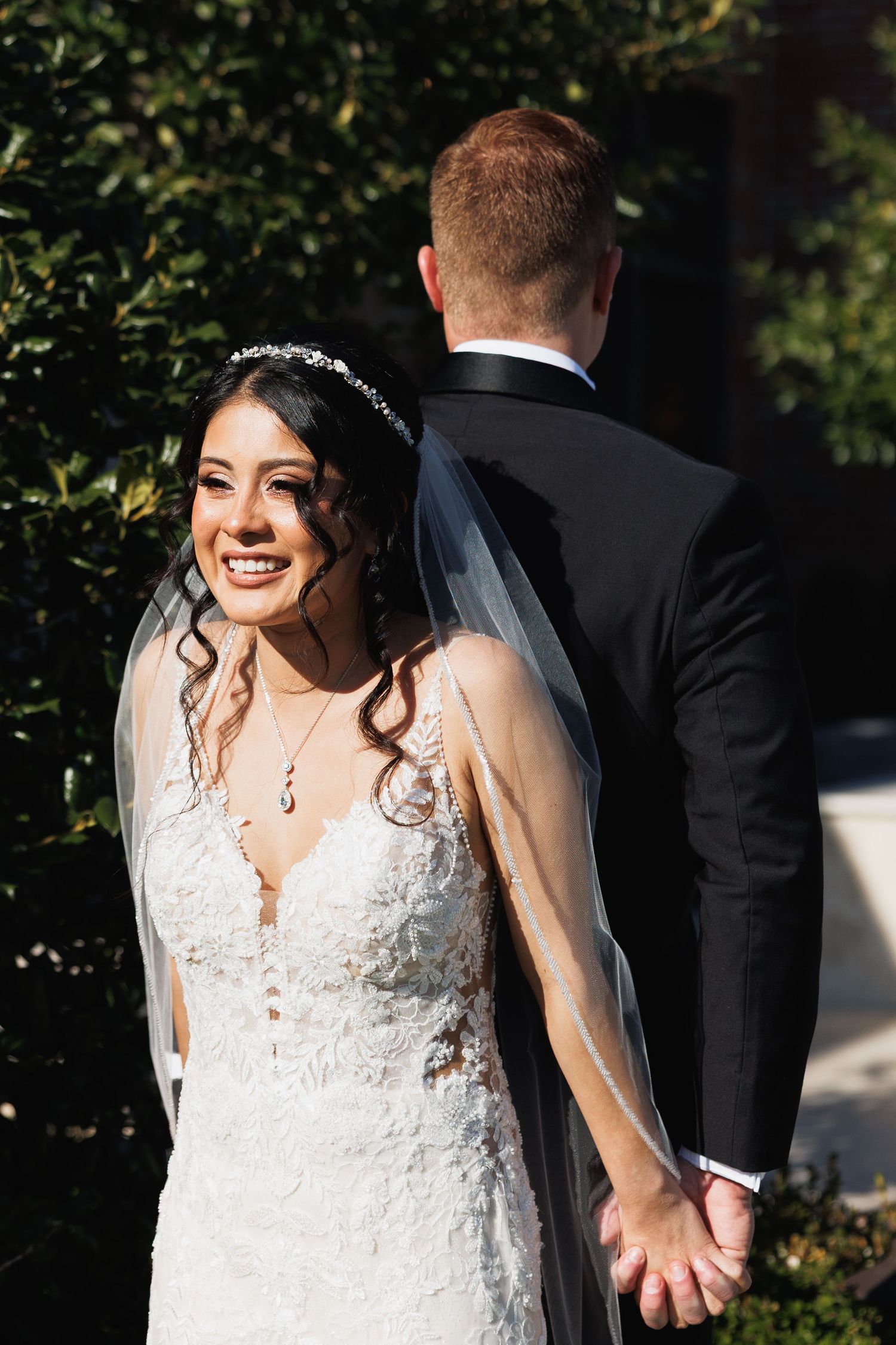 Radiant bride in lace gown holds hands with groom in black tuxedo outdoors among green foliage.