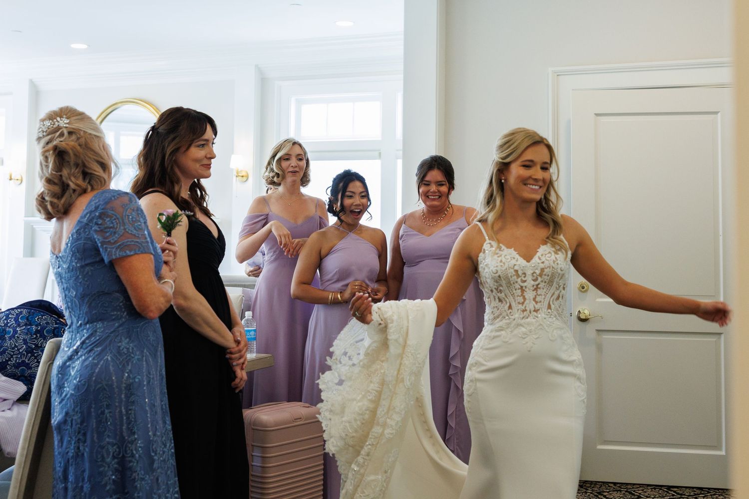 Bride in white lace gown twirls for bridesmaids in lavender dresses and family during wedding preparations indoors.