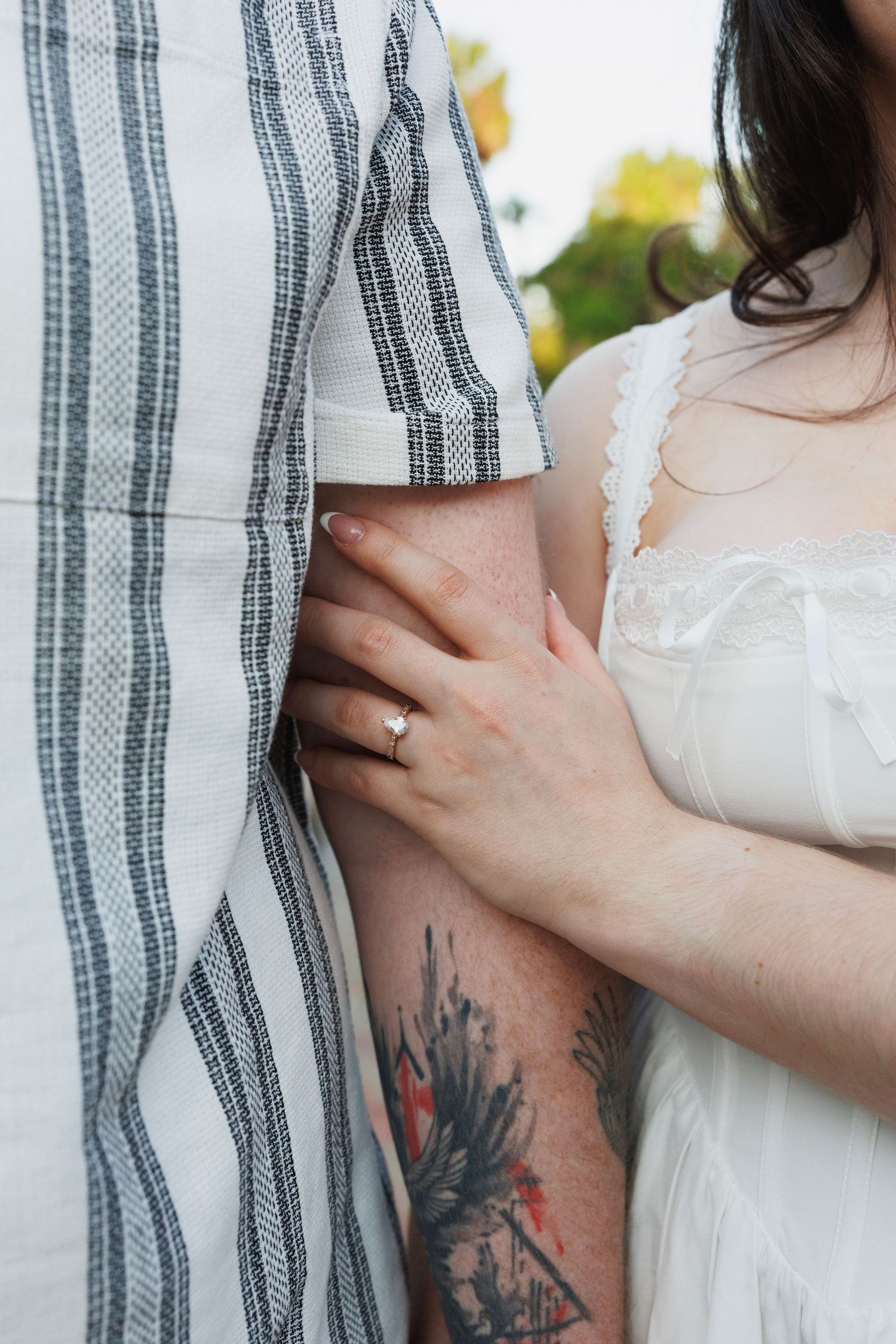 Close-up of couple embracing, woman in white dress with floral tattoo wearing ring holds man in striped shirt.