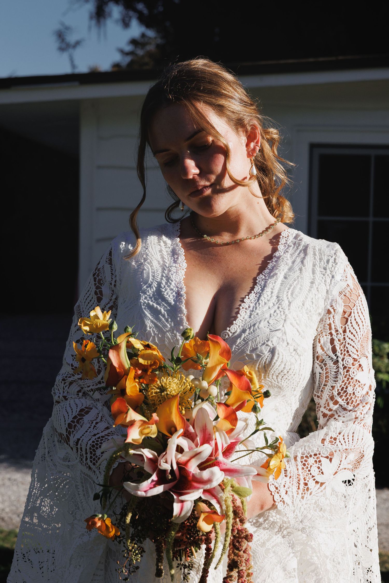 Bride in white lace boho dress holding colorful floral bouquet with lilies and orange blooms outdoors.