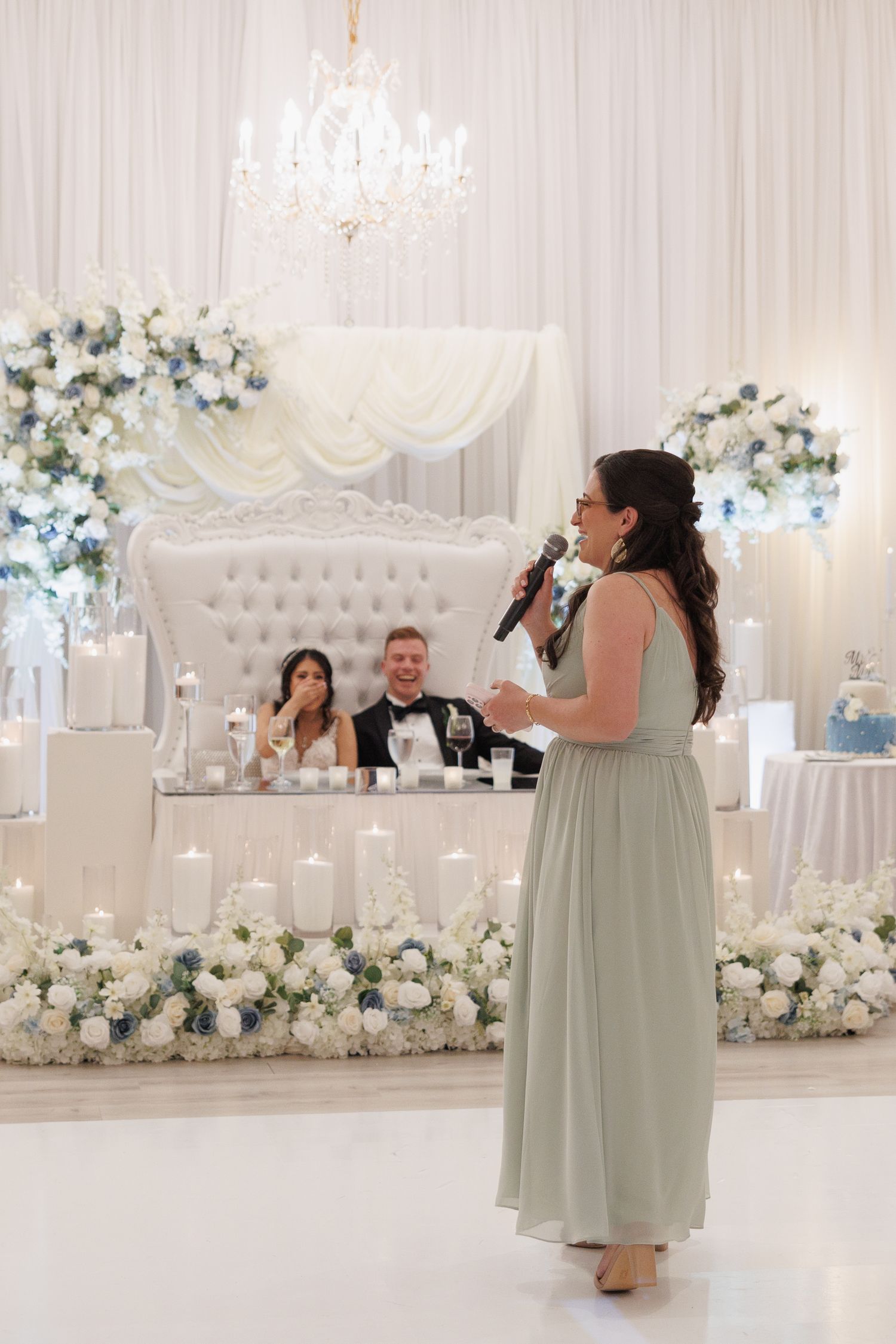 A bridesmaid in sage green gives a heartfelt speech as the bride and groom listen at their sweetheart table at the Yorkmont, NC.