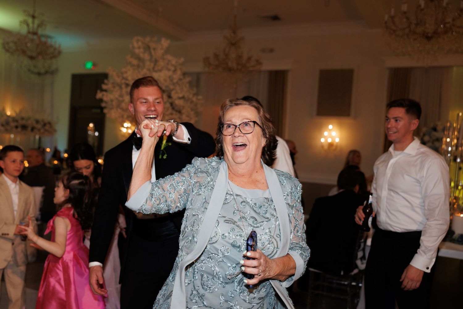 A joyful grandma dances at a wedding reception in an elegant ballroom with chandeliers and white decor at The Yorkmont in Charlotte, NC.