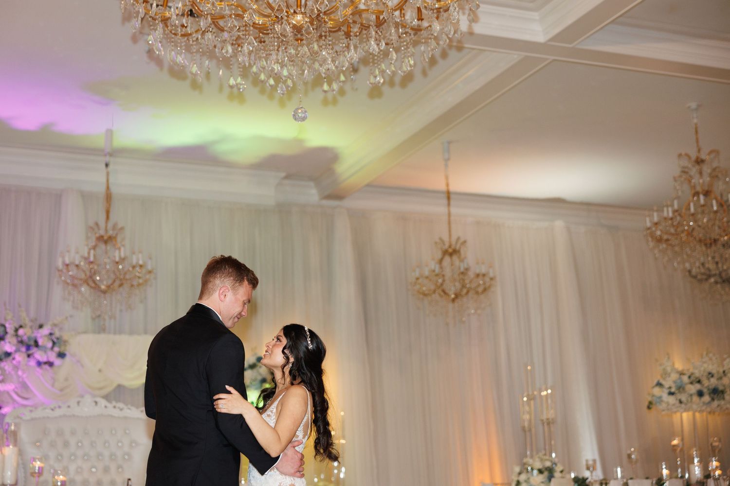 Bride and groom share an intimate first dance under sparkling crystal chandeliers in a luxurious white draped ballroom.