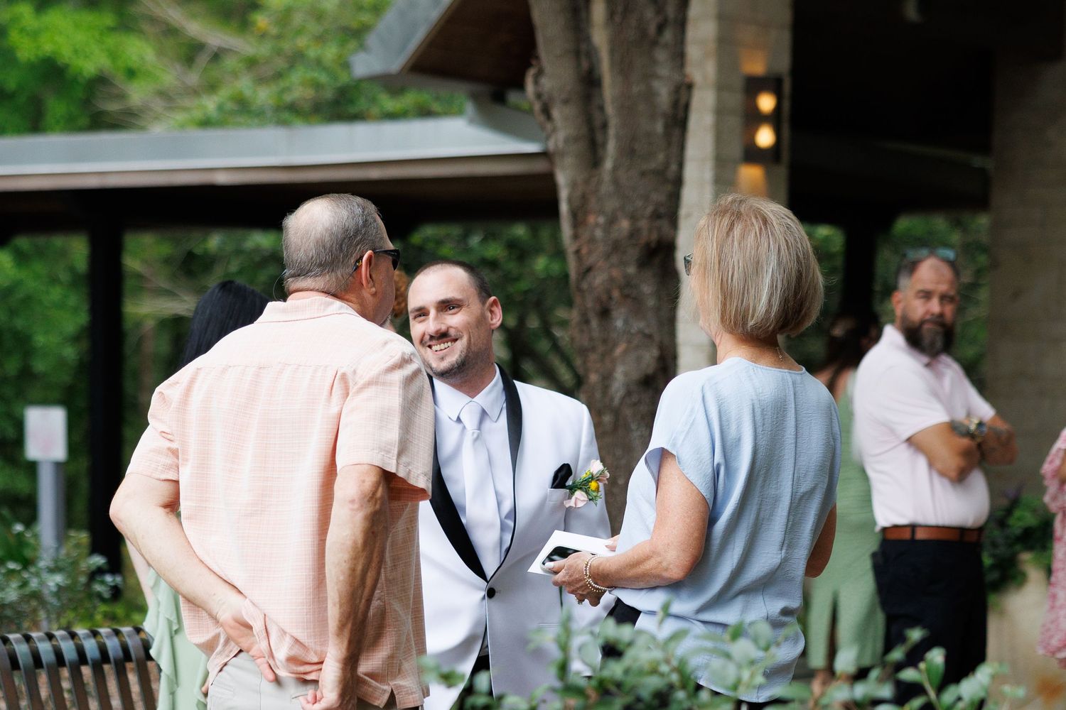 Smiling groom in white suit conversing with wedding guests outdoors near lush greenery.