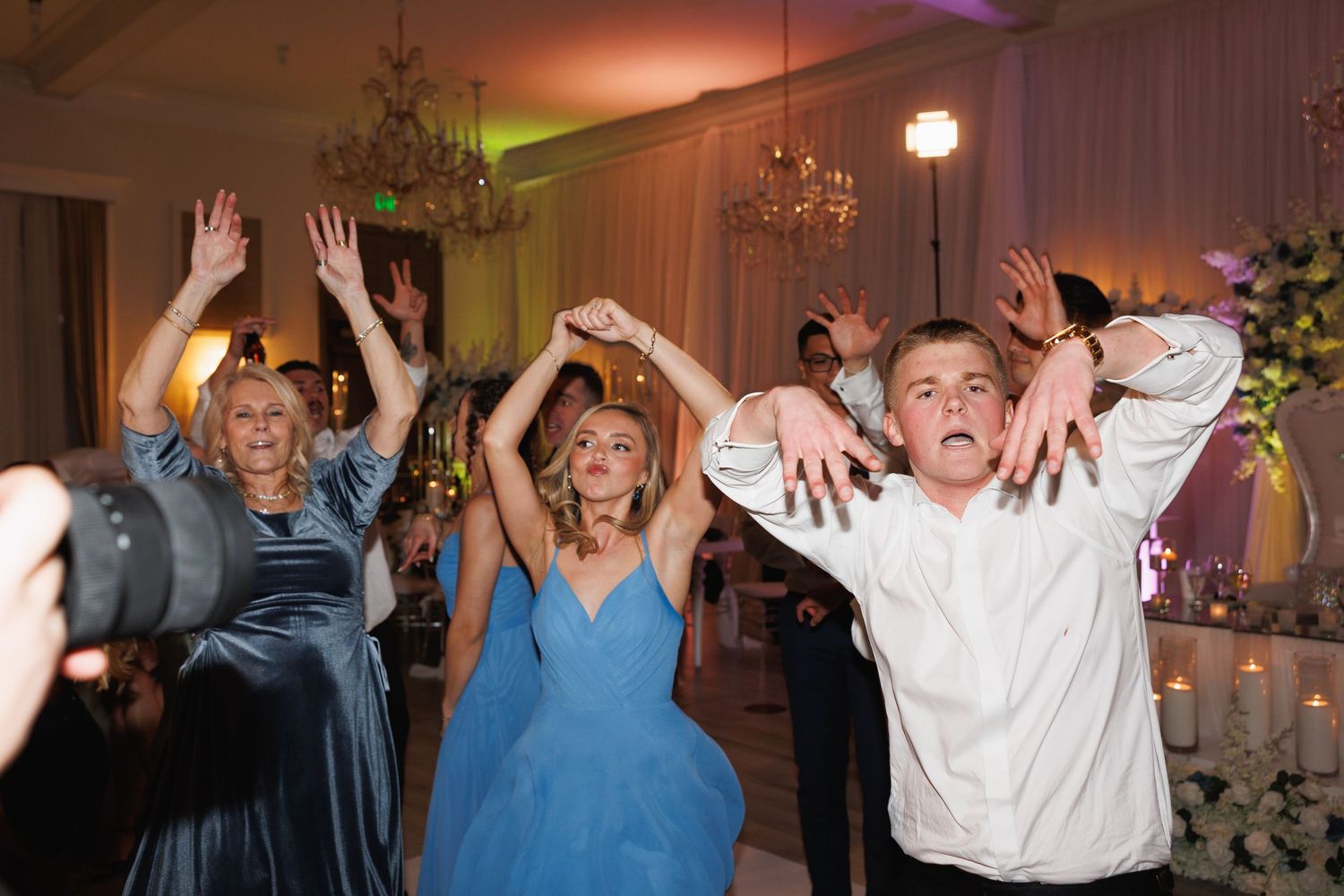 Wedding guests raise their hands and dance energetically at a reception in a decorated ballroom.
