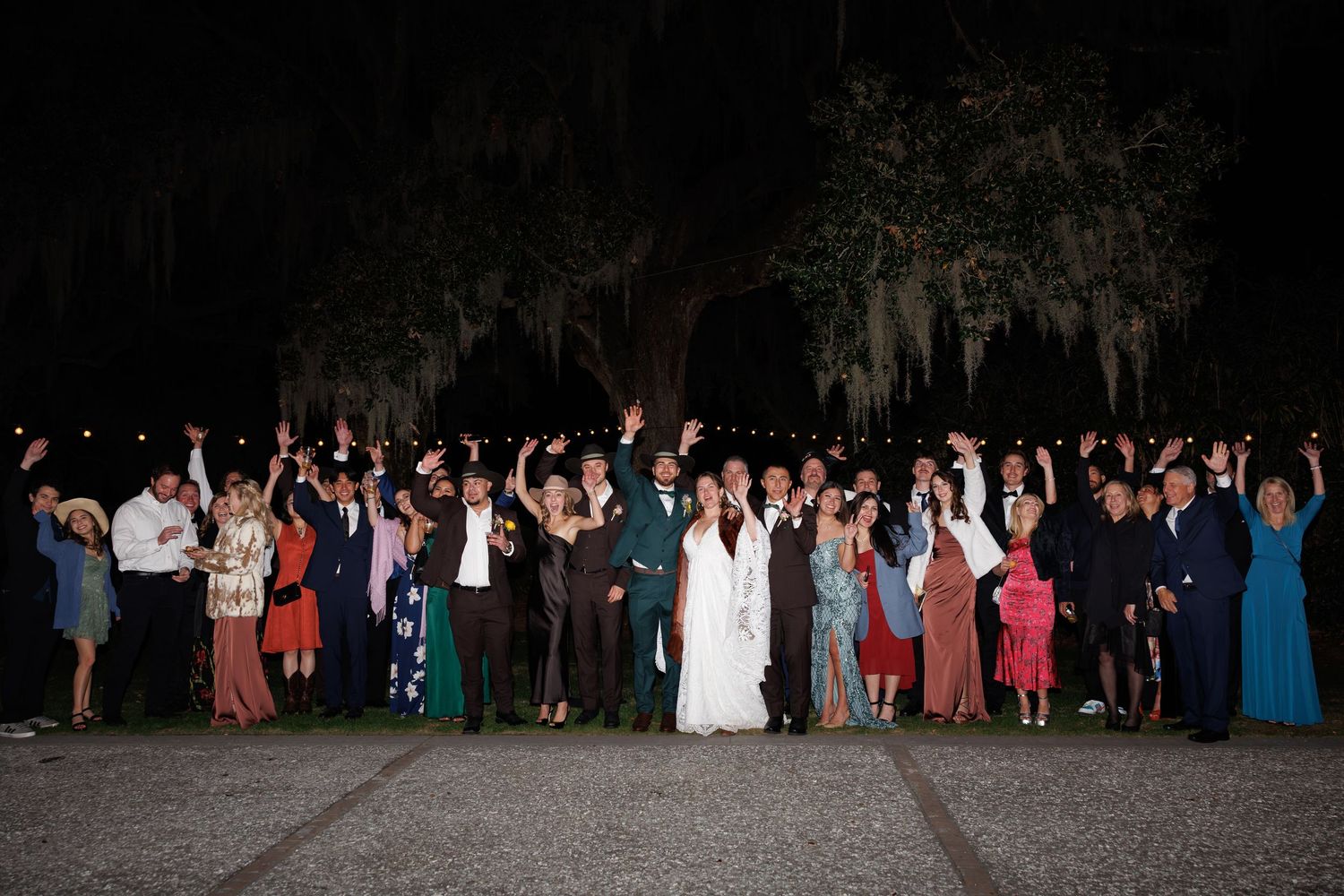 Wedding party and guests raise hands in celebration under Spanish moss trees at night reception.