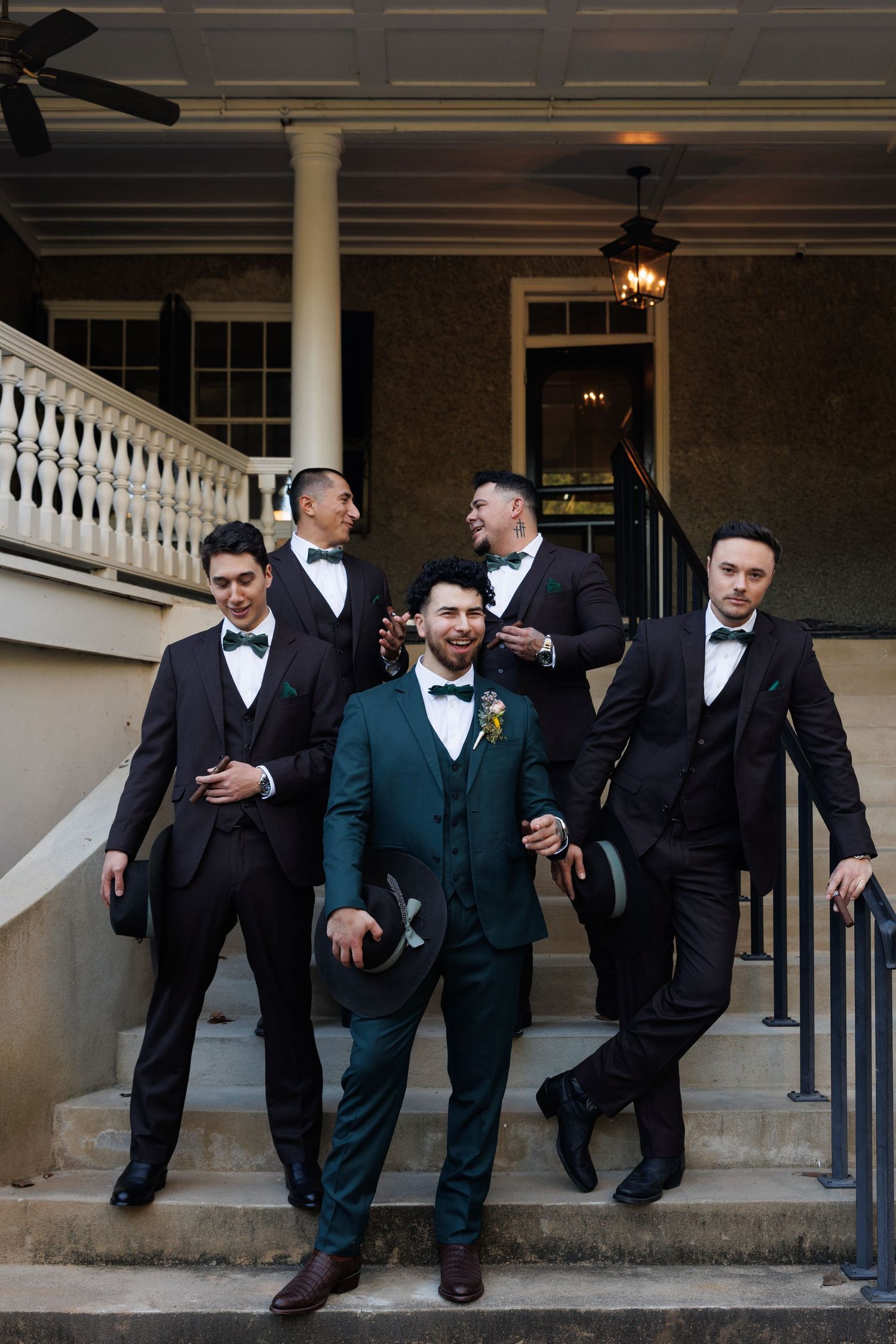 Groom in teal suit with four groomsmen in dark suits and bow ties posing on mansion steps.