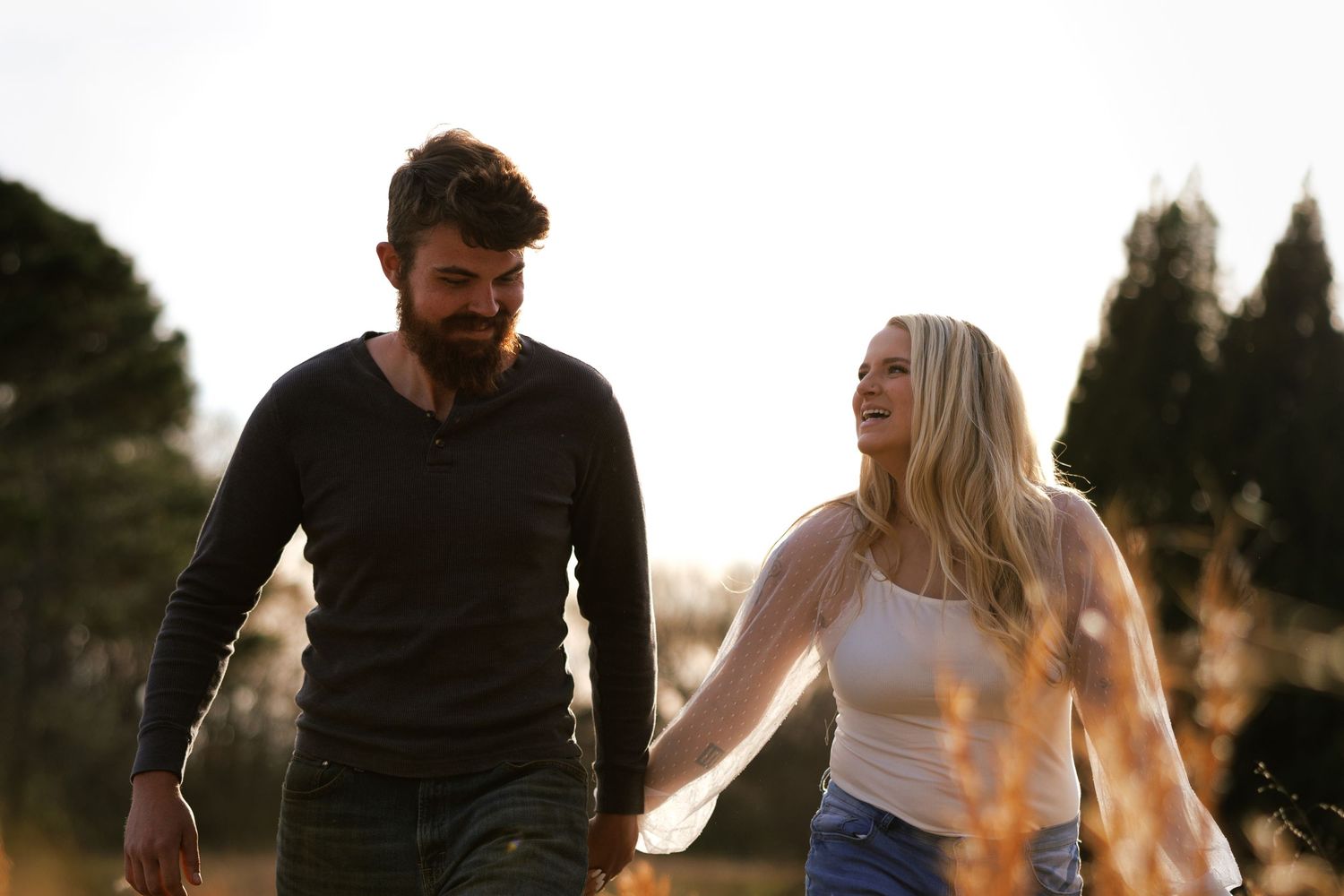 A bearded man and blonde woman hold hands and laugh while walking outdoors in golden hour light.