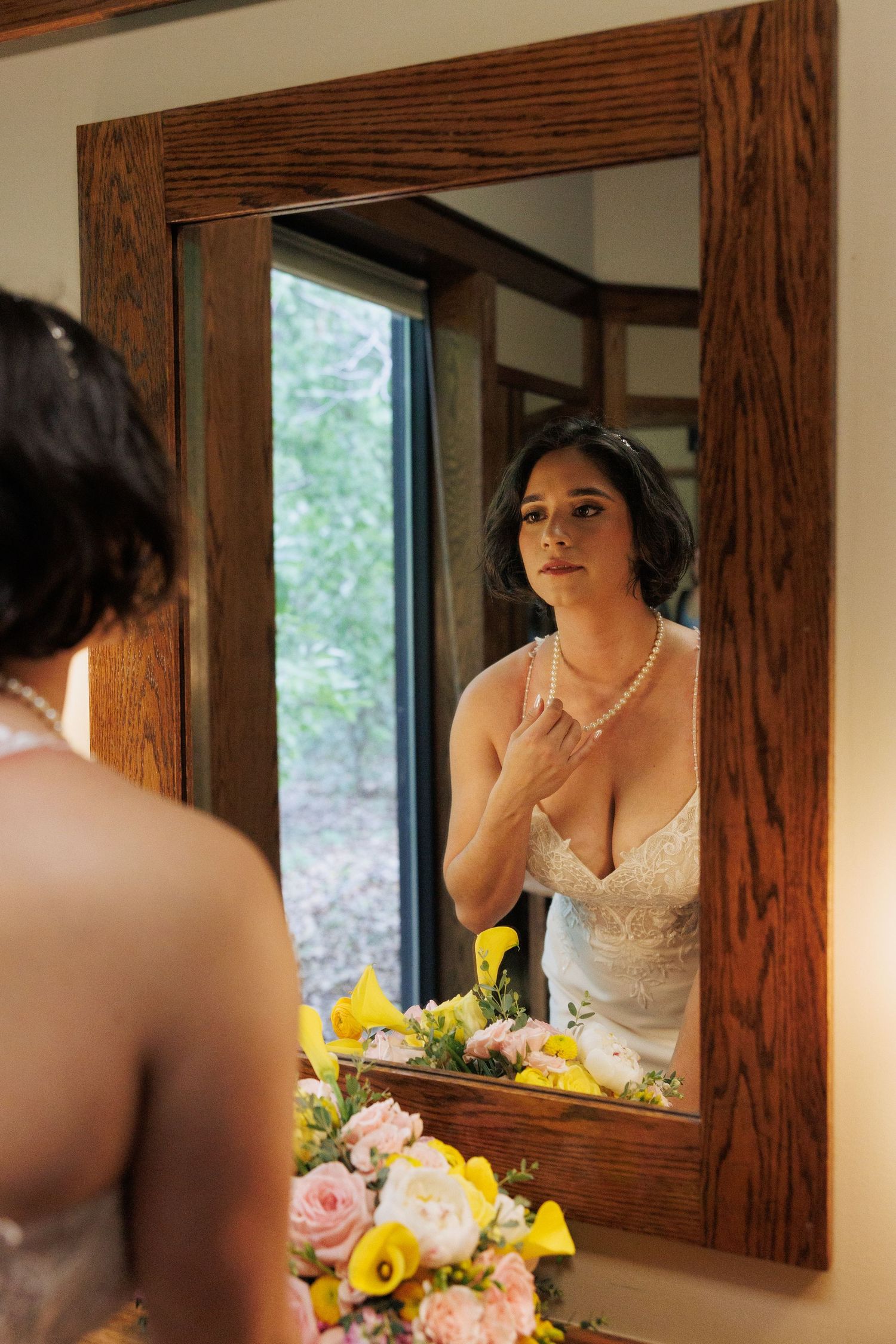 Bride in lace wedding dress adjusting pearl necklace while looking in wooden framed mirror.