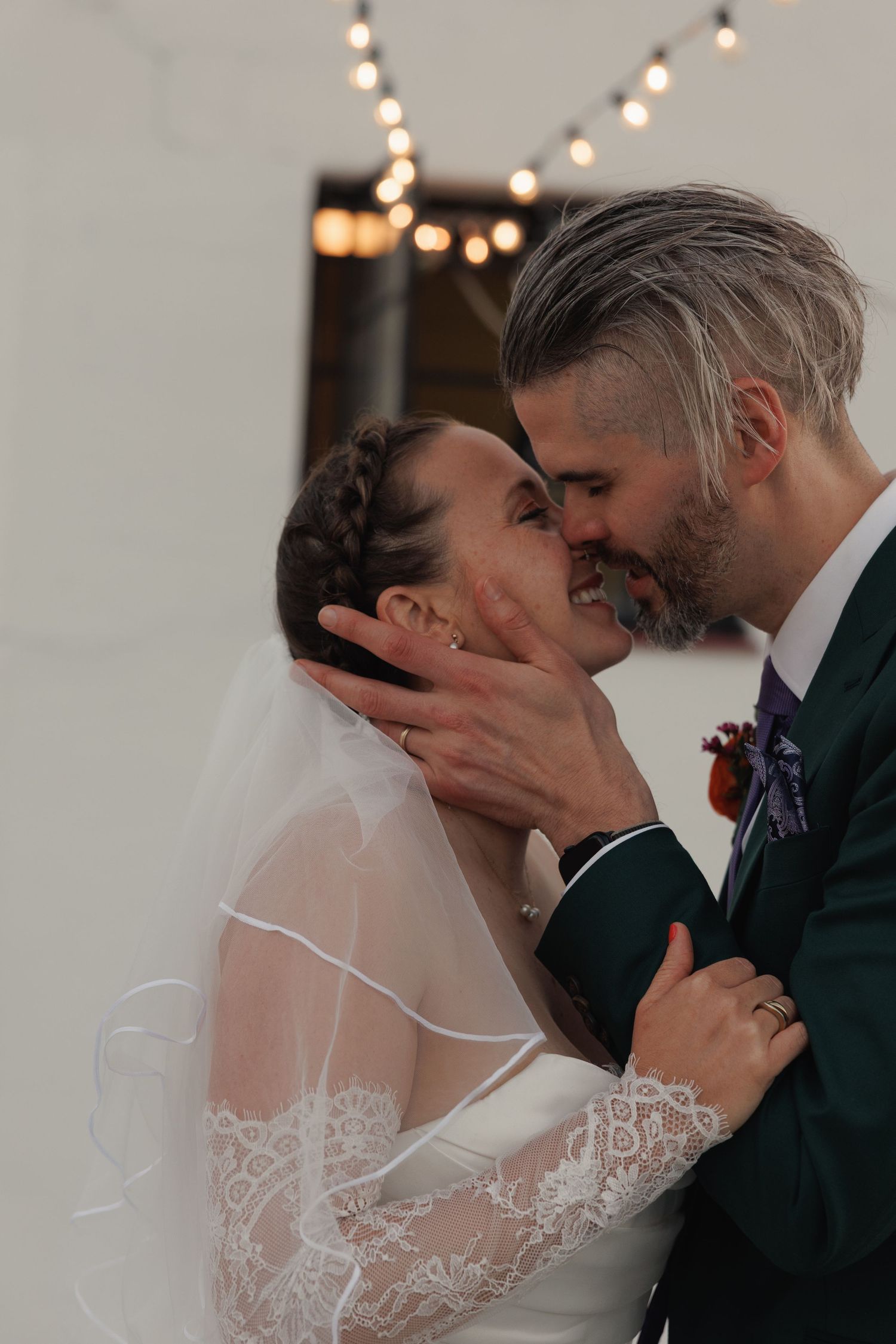 A bride and groom share their first kiss, with the bride wearing a lace veil under warm string lights.