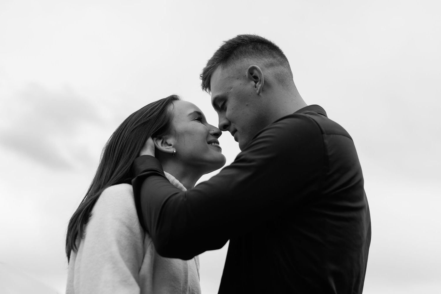 Black and white photo of a romantic couple sharing an intimate moment, almost kissing outdoors against a cloudy sky.